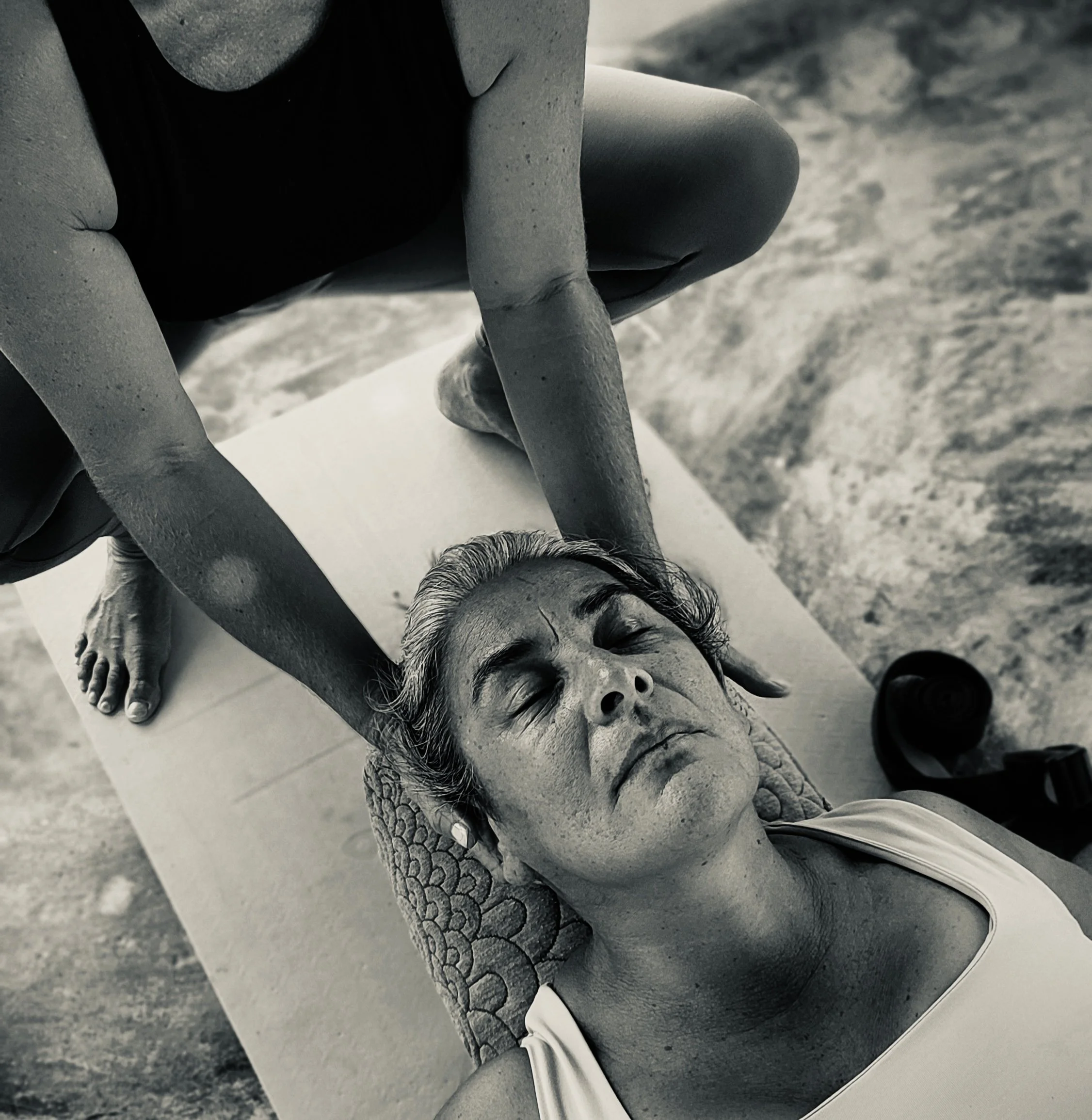 Close up of a woman reclined on her back in a yoga class, eyes closed. She looks relaxed. Another woman is partially in the frame, her hands are cradling the first woman's head in a comforting hands-on adjustment.