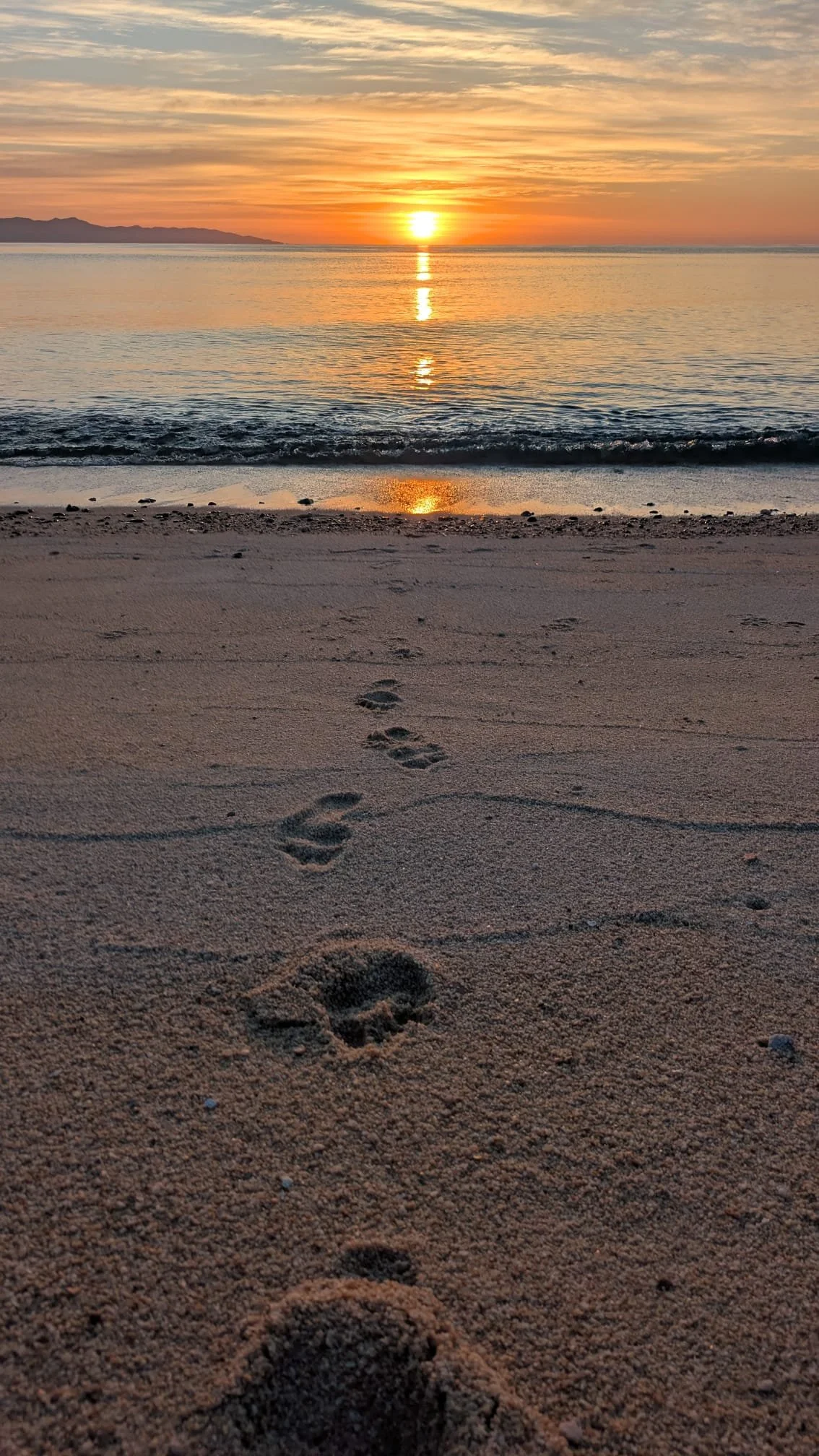 Footprints in the sand on the beach during sunset, with calm ocean waves and a distant mountain range.