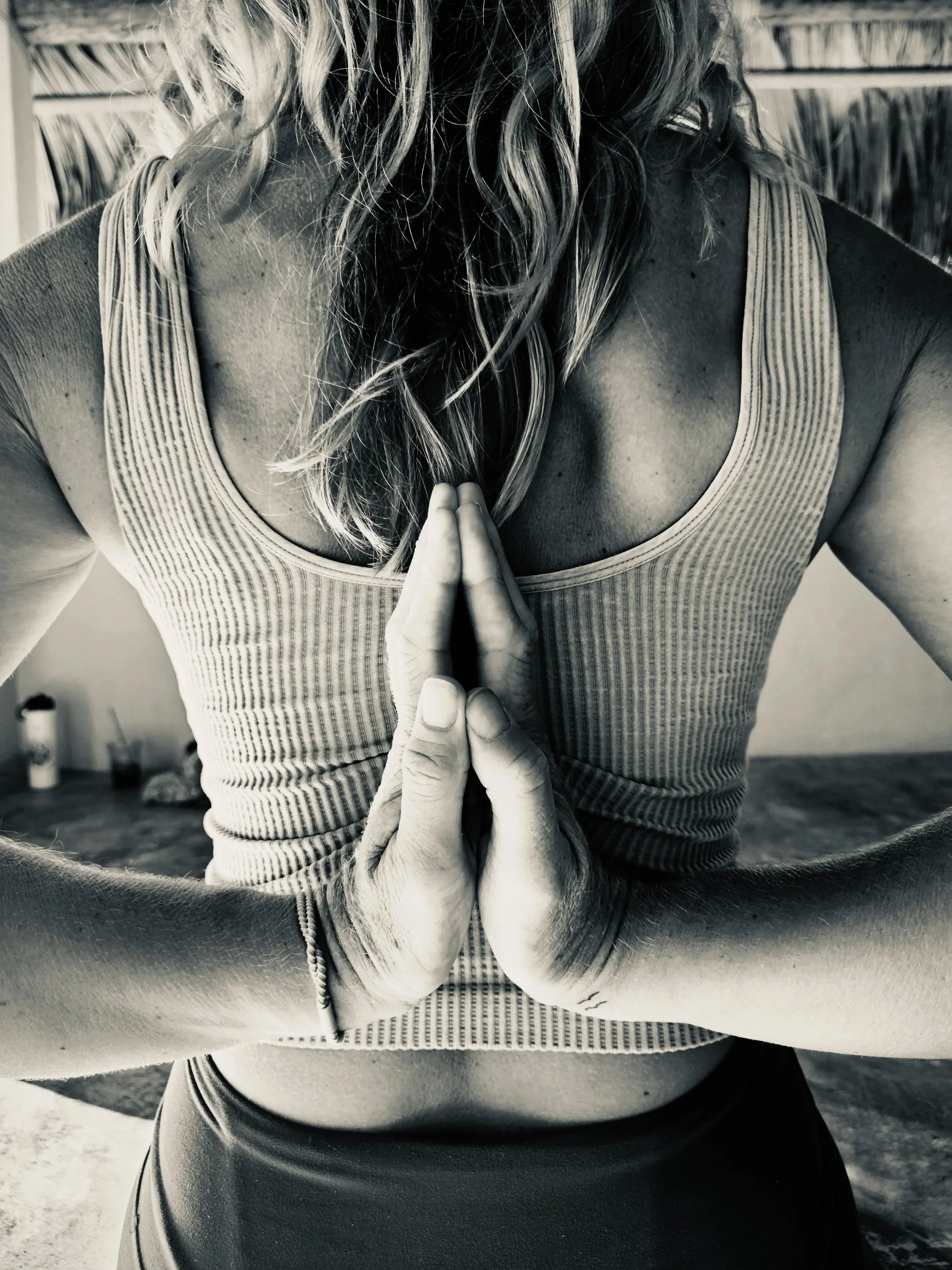 A person with wavy hair and a striped tank top is practicing yoga, with their hands pressed together in a reverse prayer position behind their back.