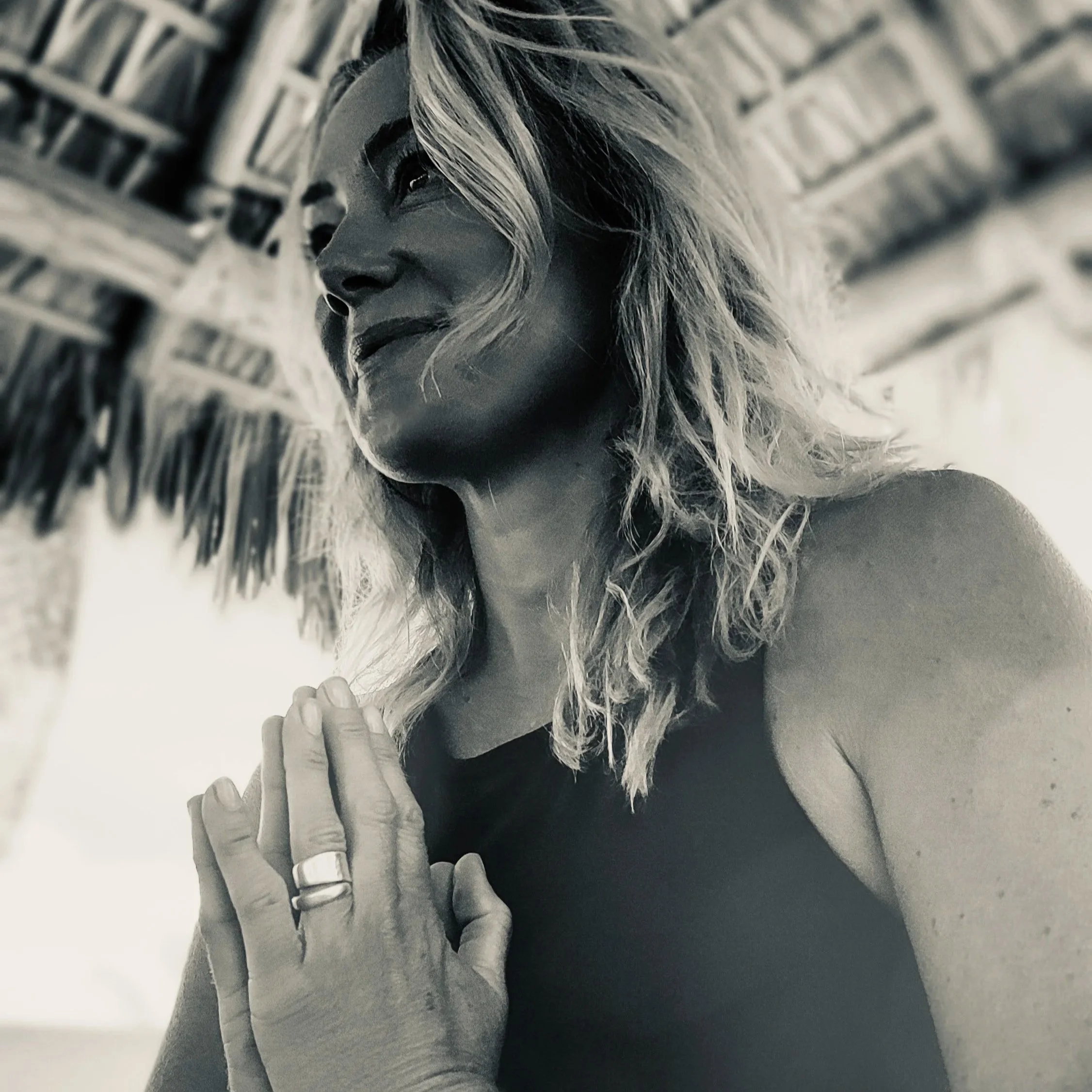 Close-up of a person's face with long, wavy hair covering part of the face, looking to the side, with a blurred background. Hands in prayer/namaste in front of her chest.