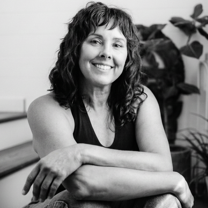 Black and white photo of Valerie Moselle. Her hair is curly hair, she is wearing a sleeveless top and sitting indoors on steps with a plant in the background.