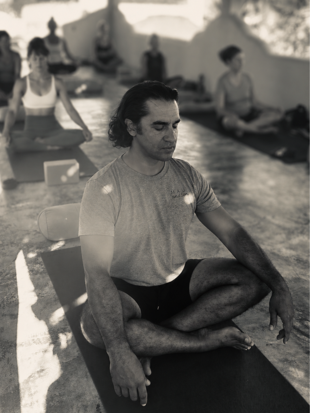 A man practicing yoga in a seated cross-legged on a yoga mat in an outdoor setting, with several other people yoga practicing in the background.