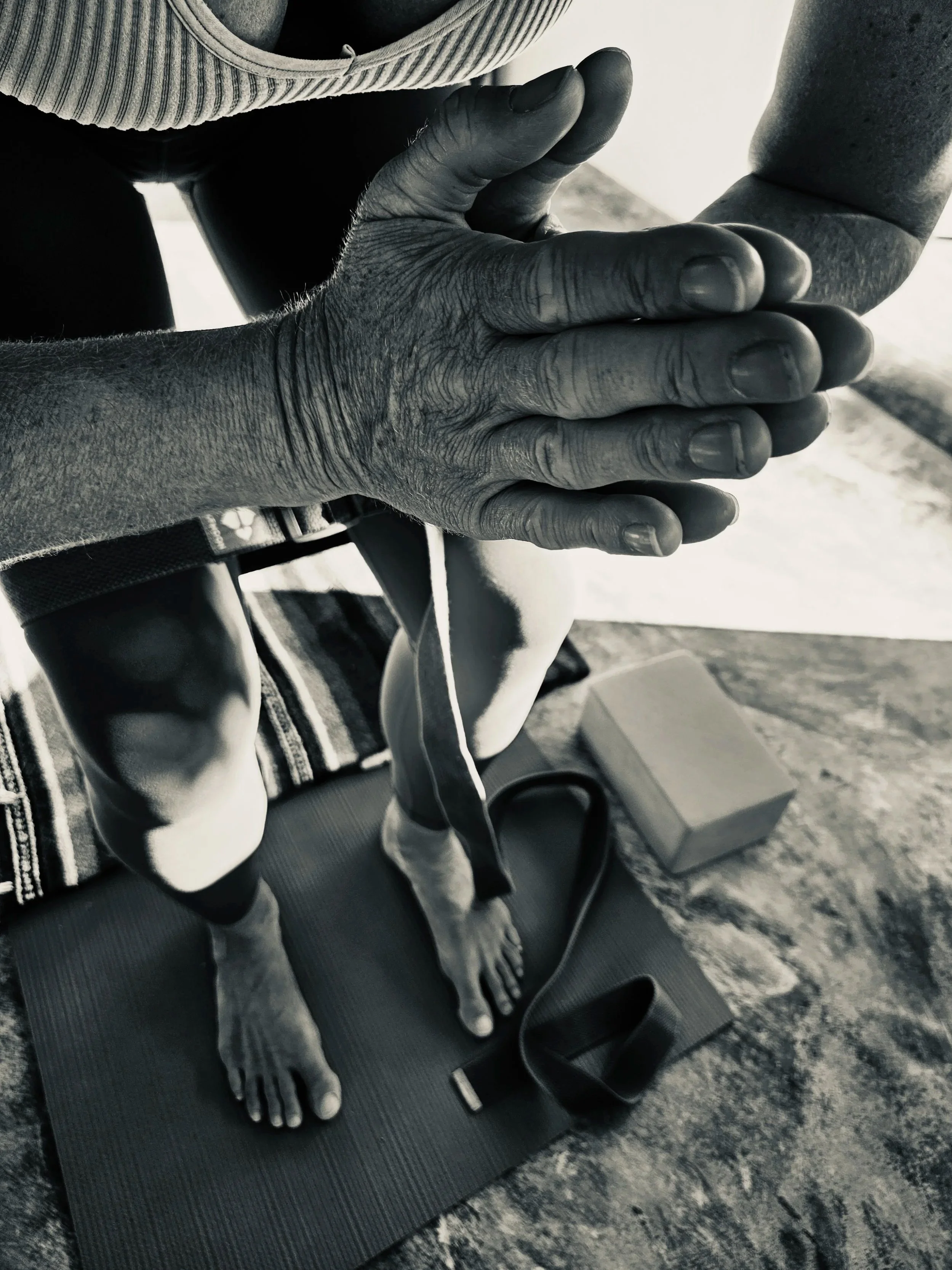 Close-up of a person's hands in prayer at her breastbone. She is practicng chair pose on a black yoga mat, with a water bottle, and a yoga block on the ground near her. The image is taken from above, so you can see her feet underneath her.