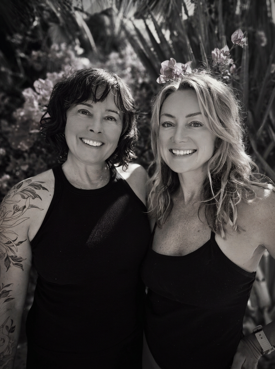 Two women smiling outdoors, one with short dark hair and tattoos on her arm, and the other with long wavy blonde hair in front of a floral background, both wearing black tank tops.