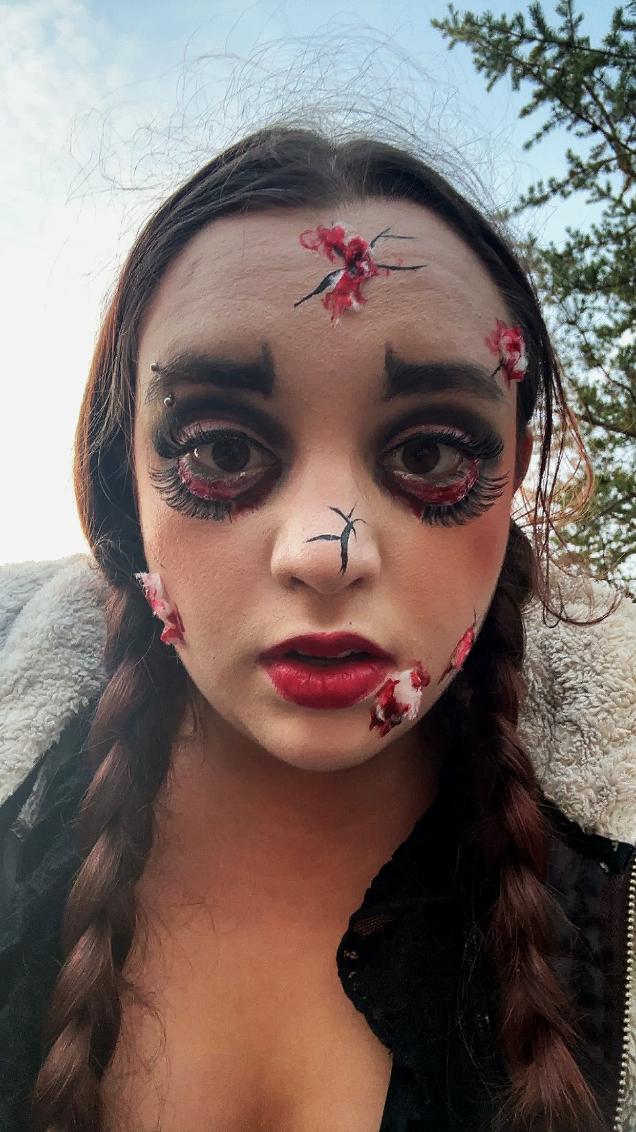 A young woman with makeup resembling a horror or zombie theme, with fake wounds and blood effects on her forehead, cheeks, and chin, and long braided hair.