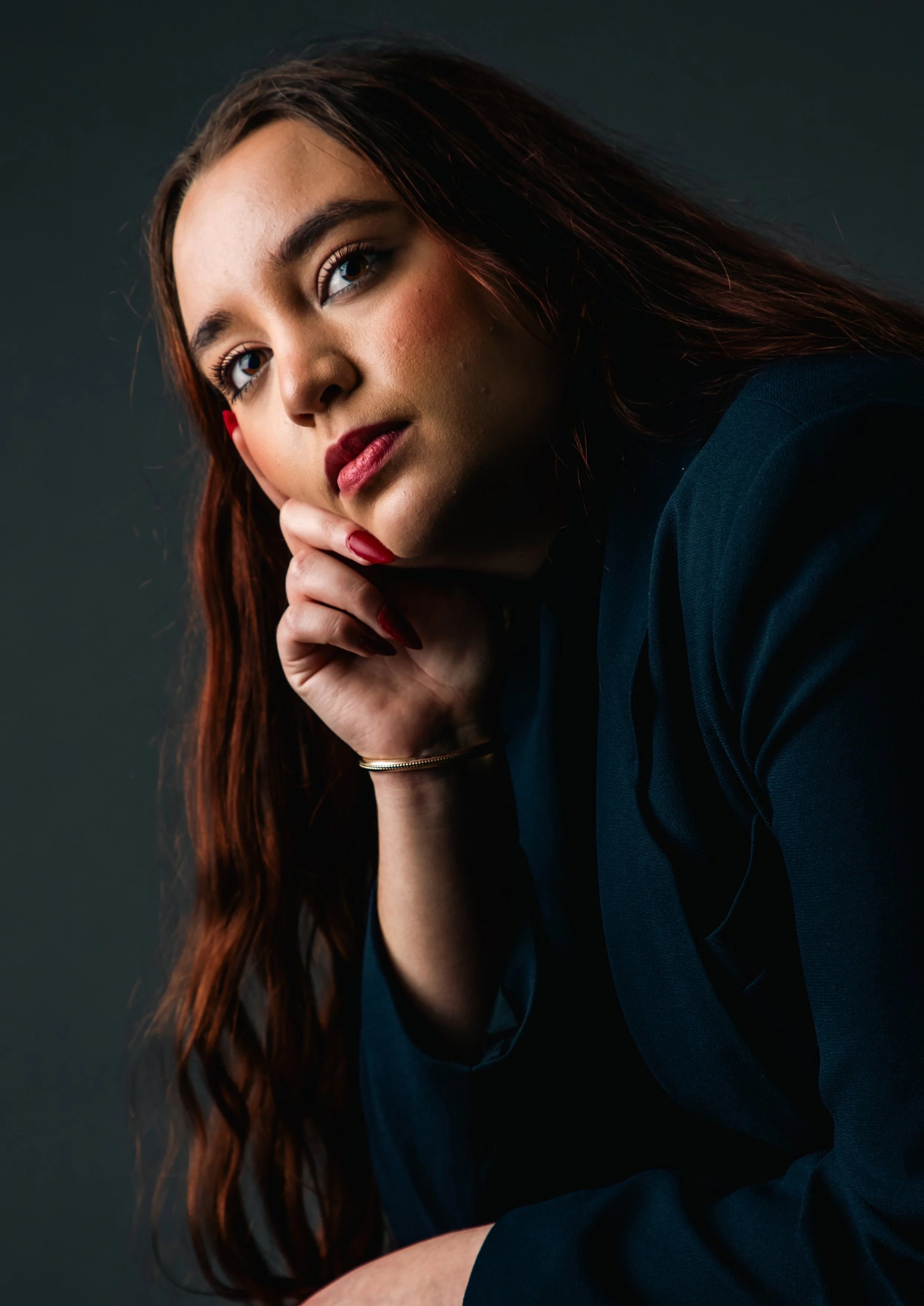 Close-up of a young woman with long auburn hair, fair skin, and dark red lipstick, resting her chin on her hand and looking at the camera against a dark background.