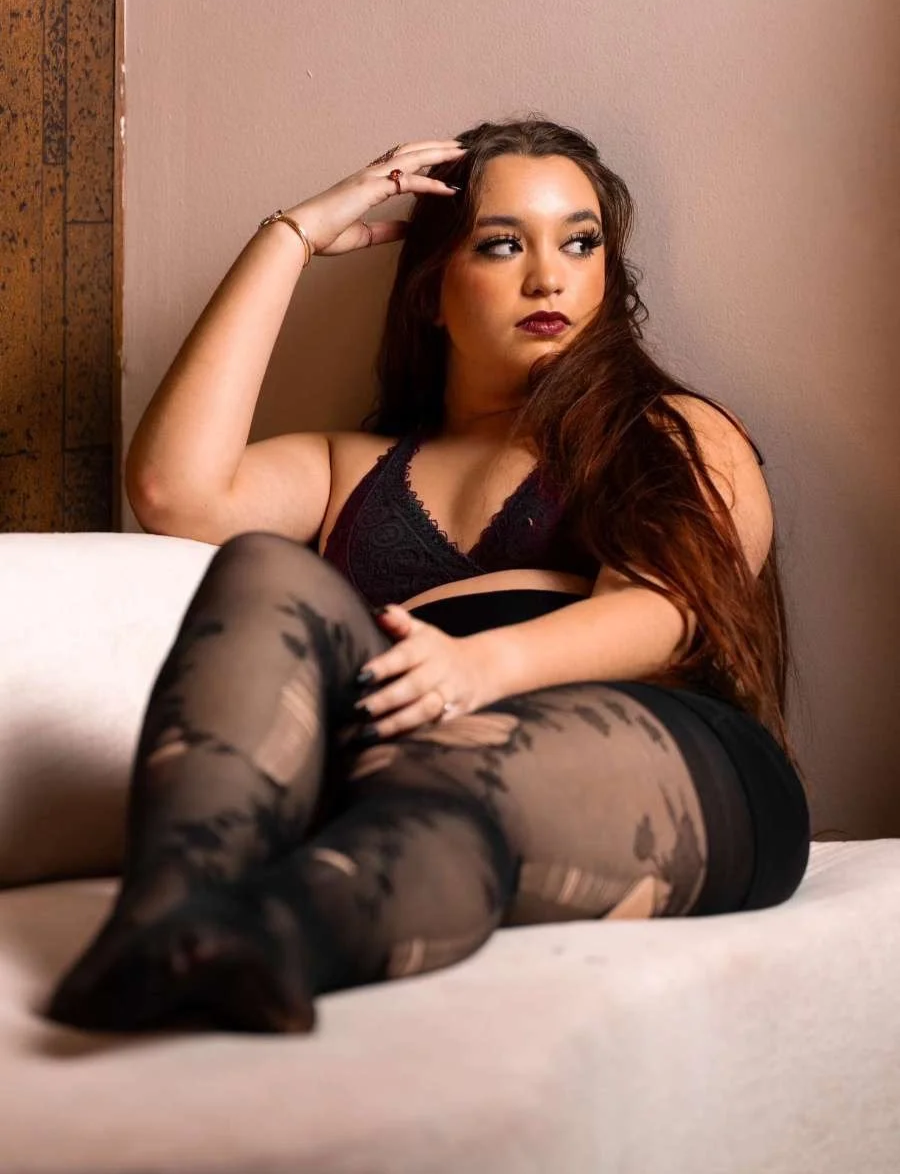 A woman with long brown hair, wearing black lace lingerie and patterned tights, sitting on a beige couch against a beige wall, looking to her right with her hand on her head.