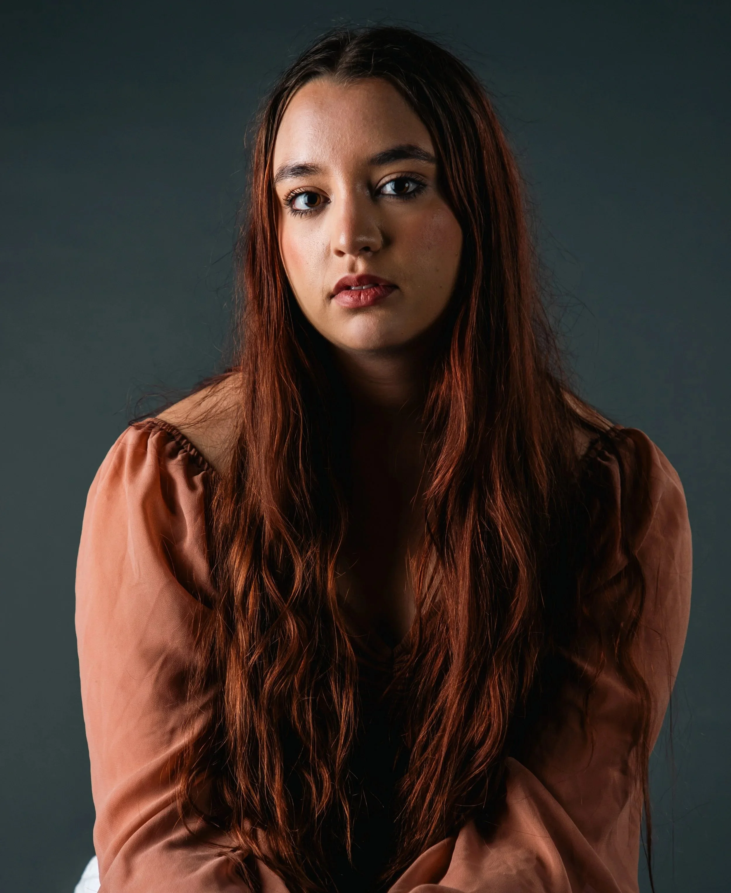 A woman with long, wavy auburn hair looking directly at the camera against a dark background.