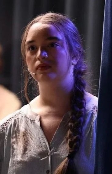 A young woman with light brown hair in a braid, wearing a light-colored blouse, standing on a stage with a dark curtain background, illuminated by a purple stage light.