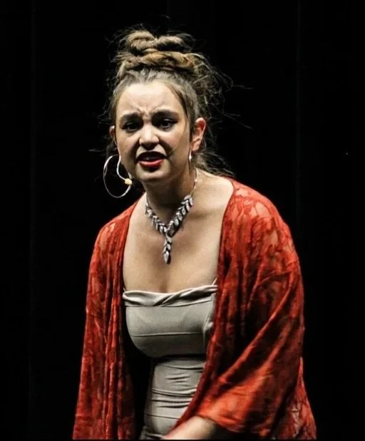 A woman with a distressed or angry expression, wearing a beige top, red patterned shawl, large hoop earrings, and a necklace, standing against a dark background.