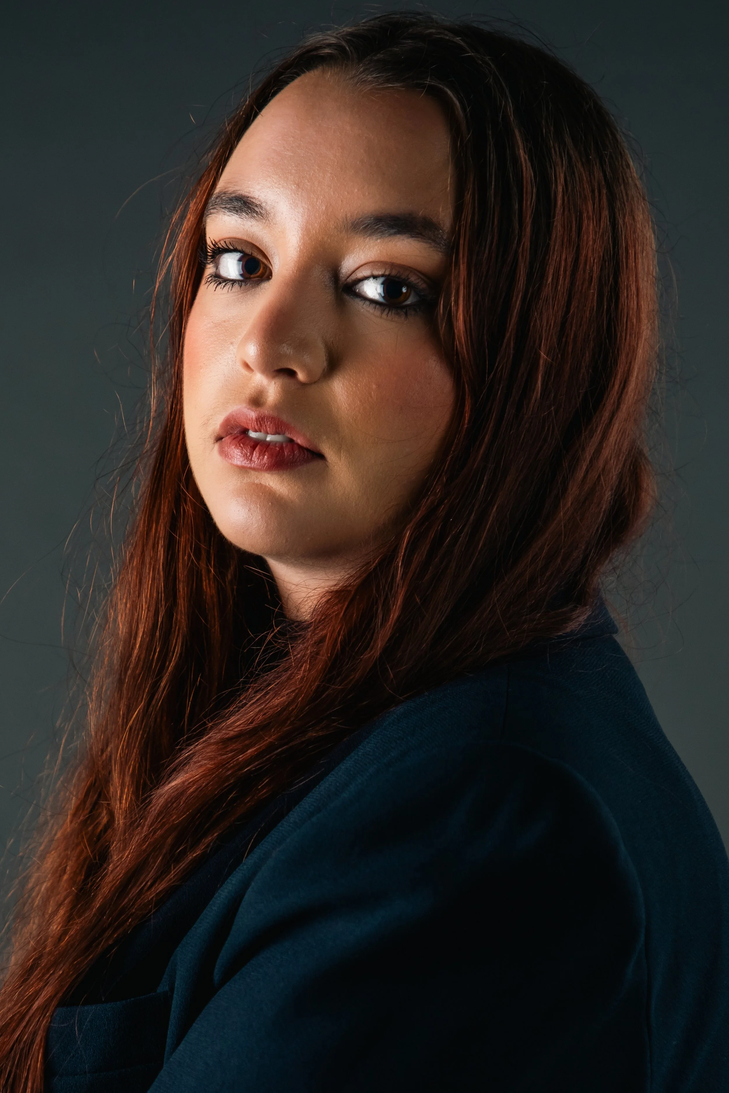 Close-up portrait of a woman with long auburn hair, wearing a dark blue top, against a dark gray background.
