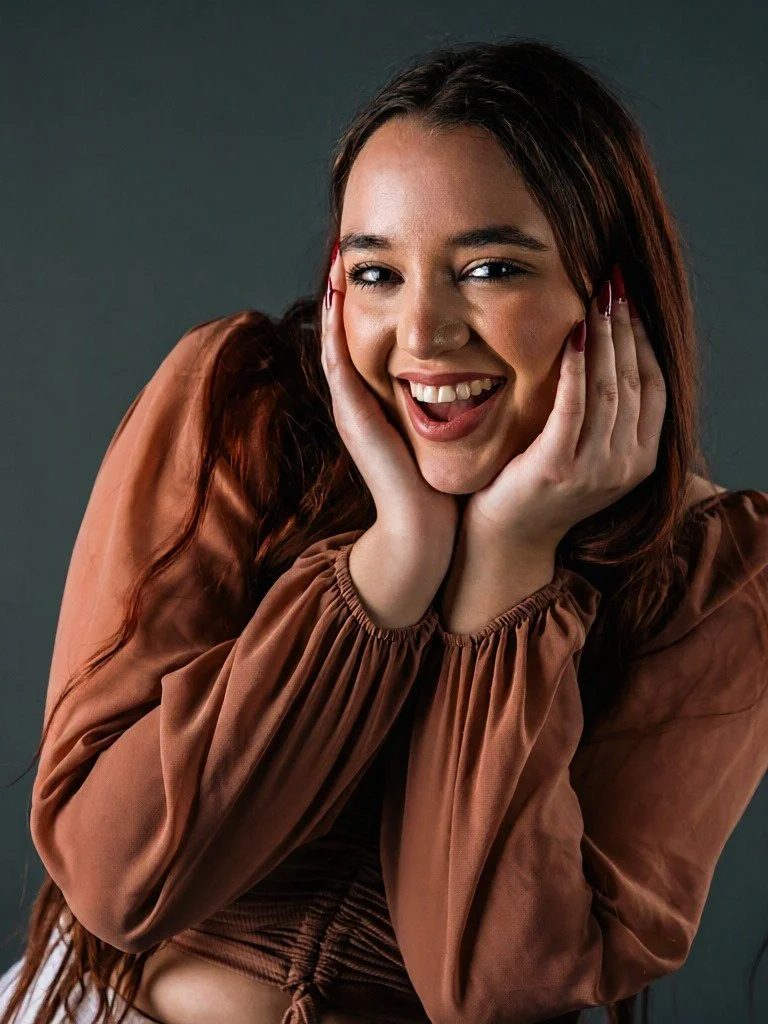 A woman with long brown hair smiling and holding her face with her hands, wearing a brown satin blouse against a dark background.