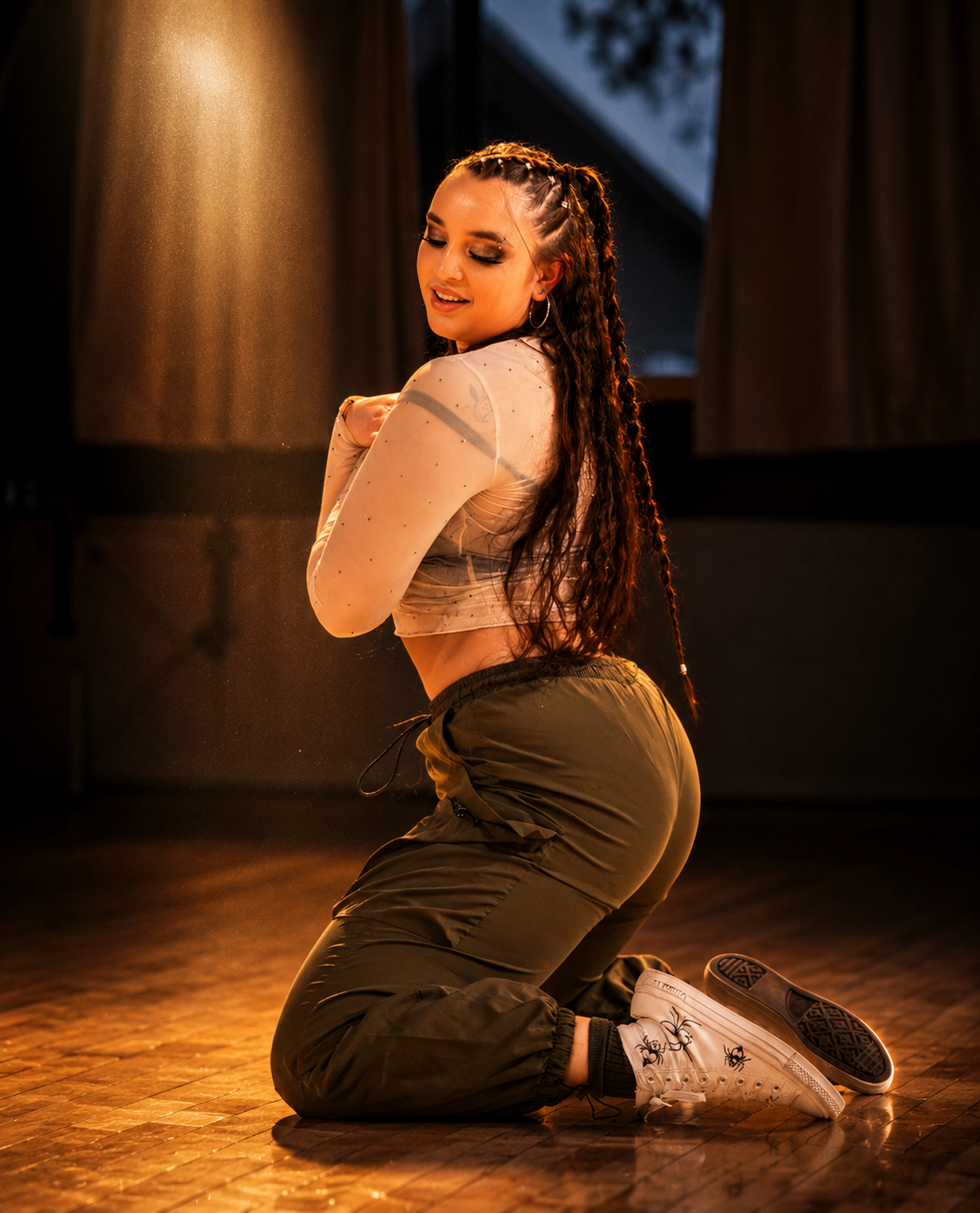 A young woman with braided hair, wearing a sheer long-sleeve top, sweatpants, and sneakers, kneeling on the floor in a dance pose with her eyes closed and a smile, in a dimly lit room with curtains and a window in the background.