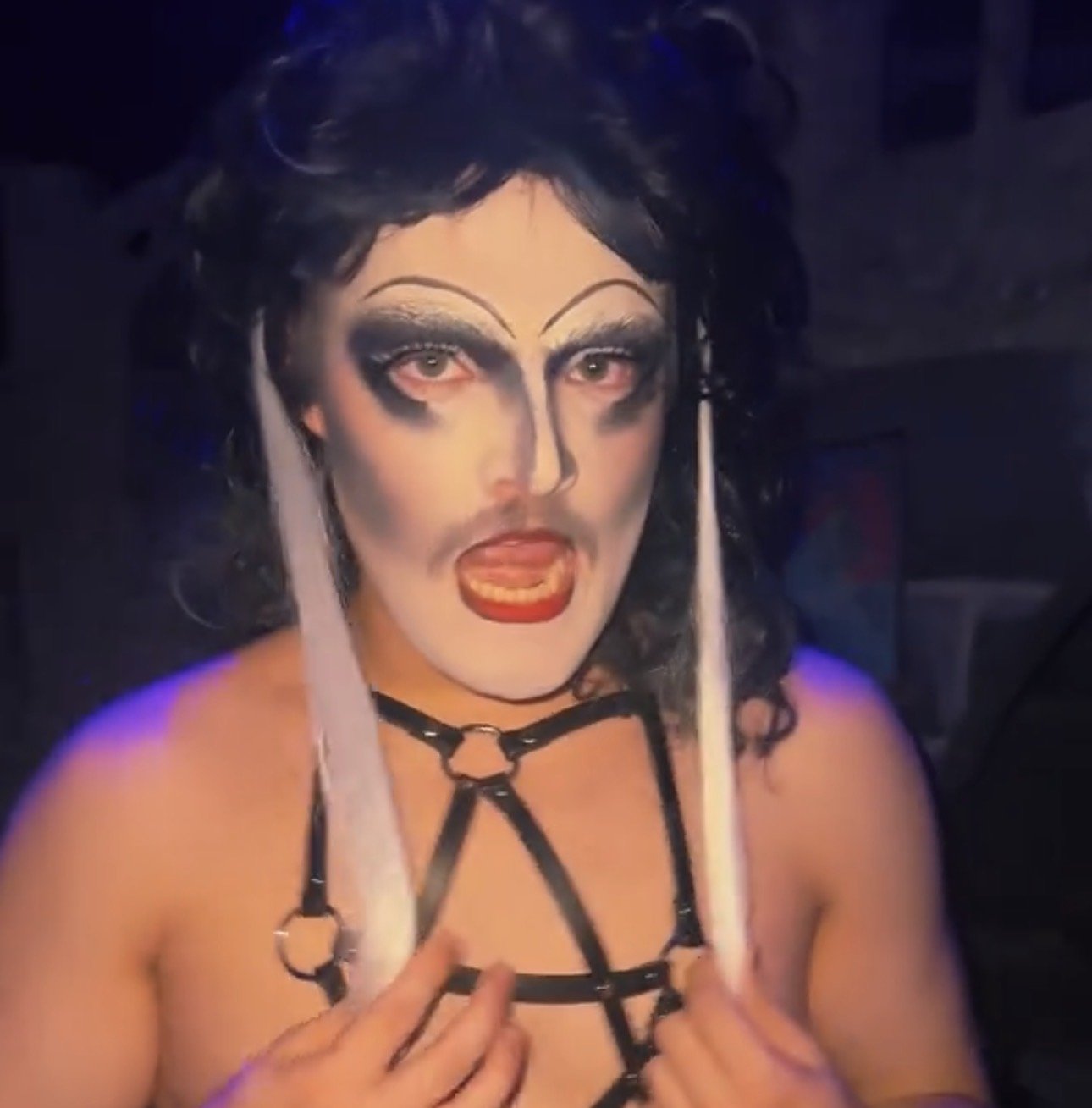 A person in drag with exaggerated makeup, black wig, leather harness, and white candles as earrings, making a provocative gesture in a dimly lit room.