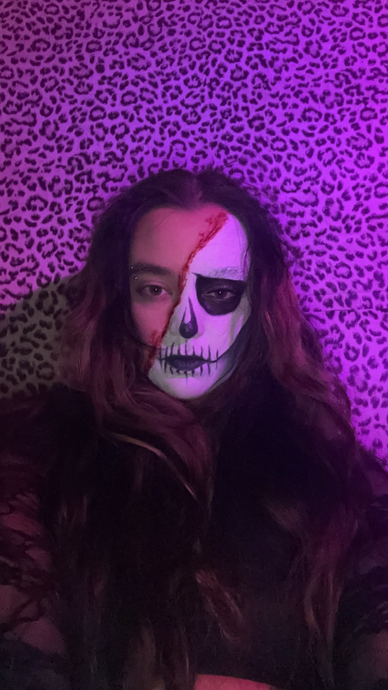 Person with skeleton face makeup, dark hair, in a dark room with purple leopard print background.