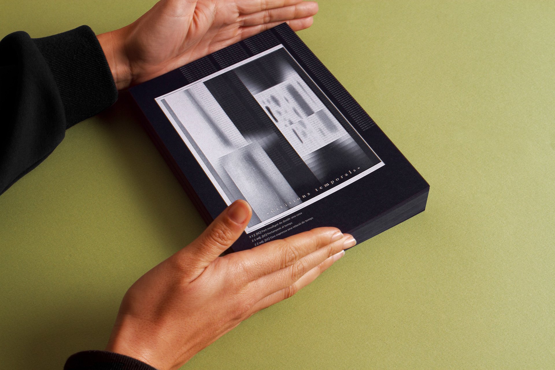Hands holding Divisions temporals, a black printed object with an abstract light study on the cover