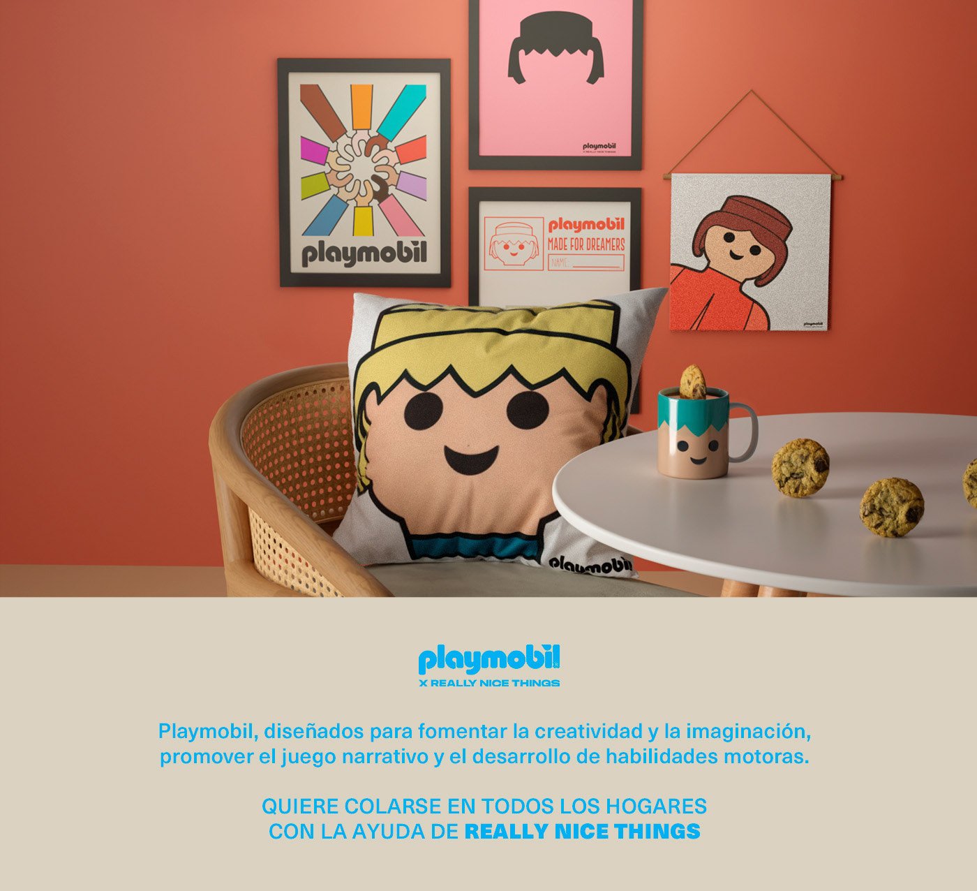 A cozy room corner with a Playmobil-themed decor. There are four framed pictures on a coral wall, including Playmobil logos and character illustrations. A cushion with a cartoon face of a Playmobil figure is on a wooden chair. A white table holds a mug with a Playmobil face and several chocolate chip cookies.