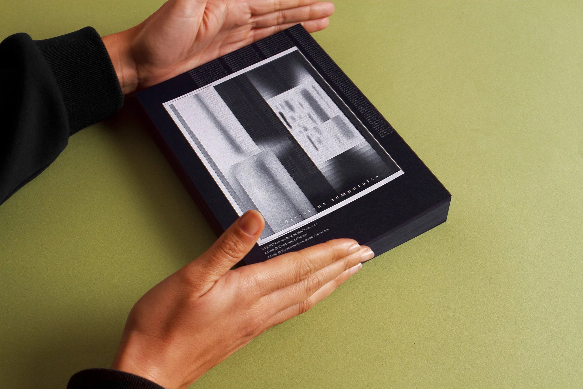 Person holding a book with both hands, revealing the bottom part of the pages, on a yellow-green surface.