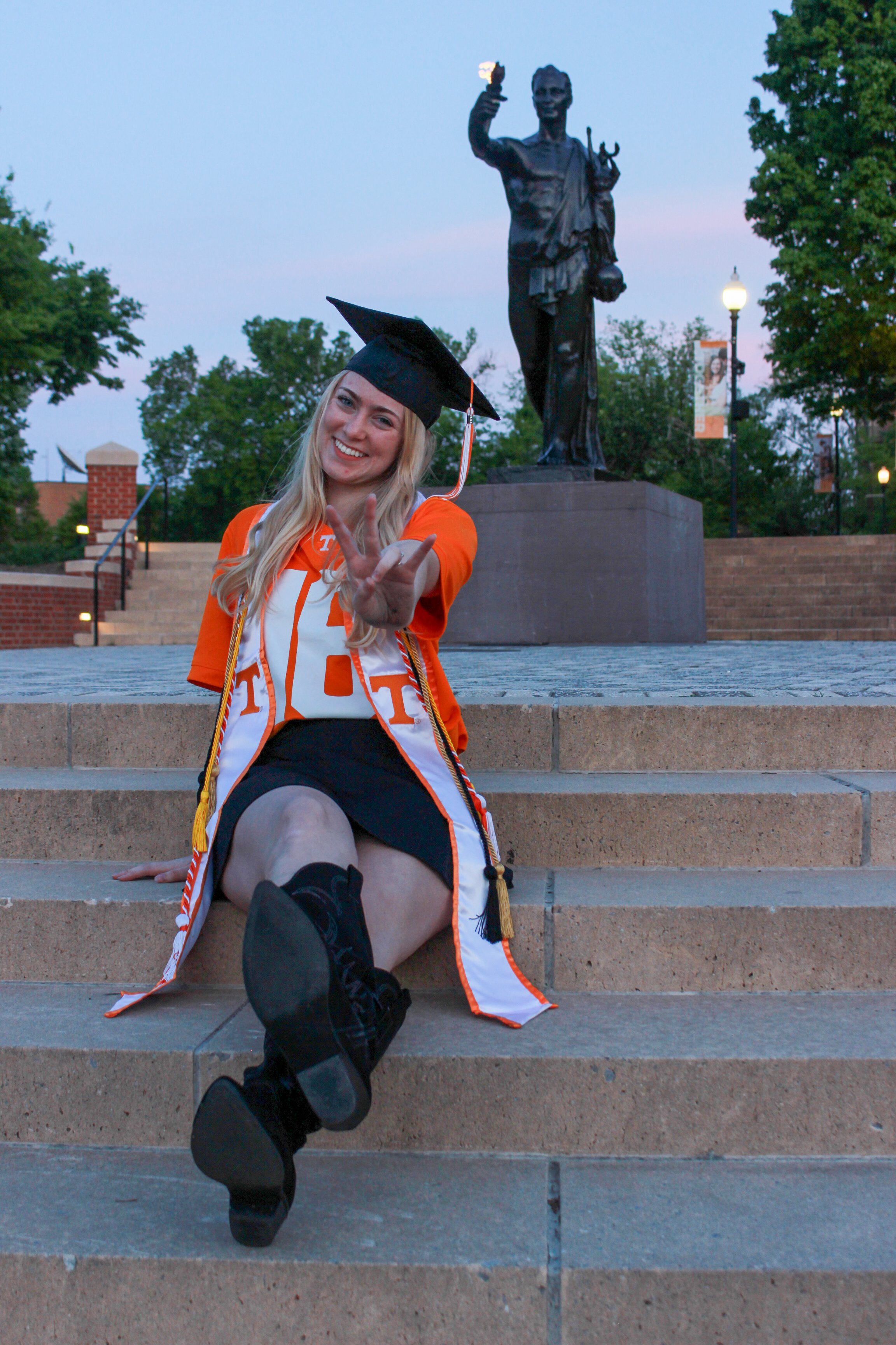 A young woman dressed in a college graduation cap and gown sitting on steps outdoors, smiling and making a peace sign with her hand, with a large statue of a woman holding a torch behind her, trees, and lamp posts in the background during dusk.