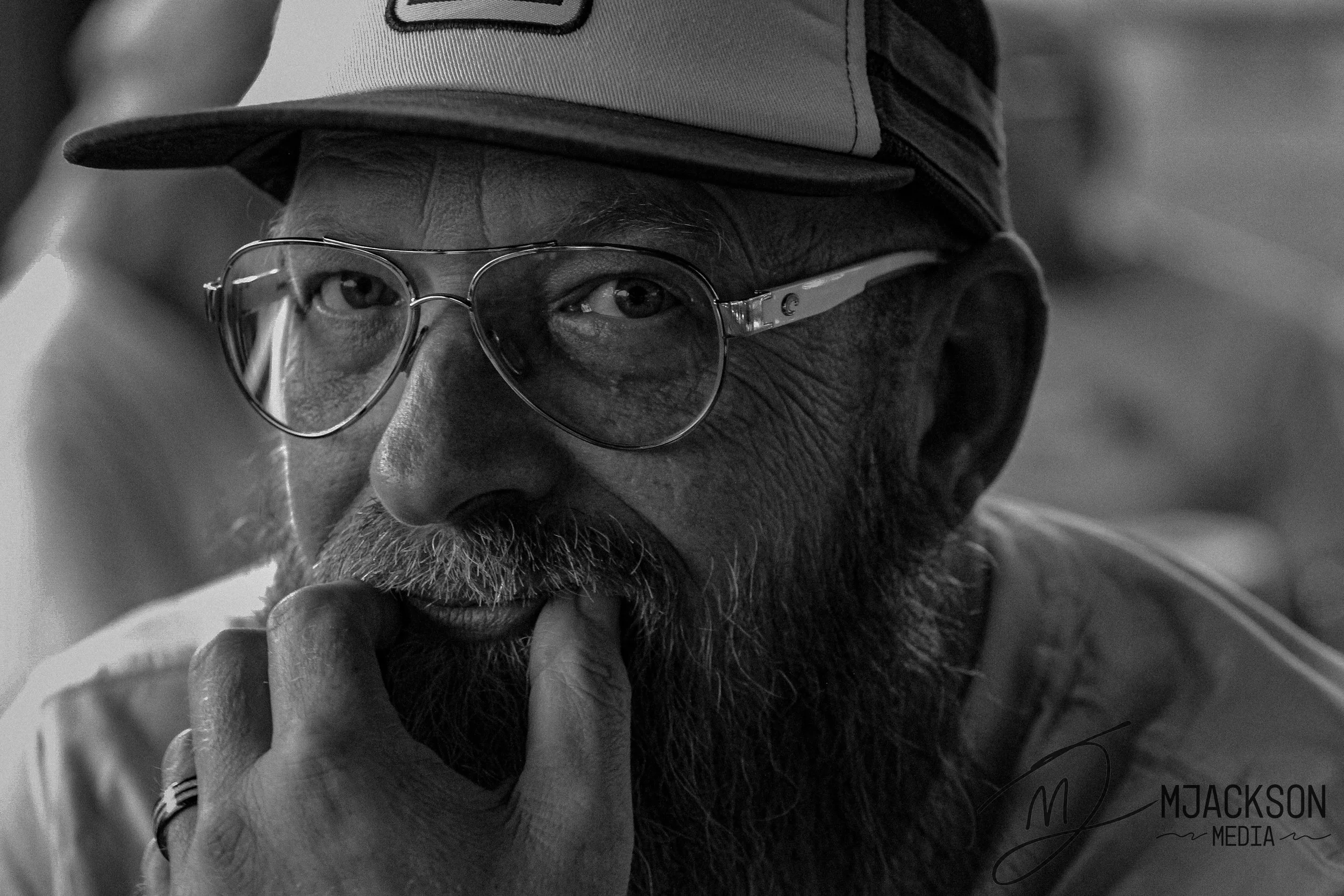 A close-up black and white image of a man with glasses, a beard, and a cap, touching his face with his fingers.