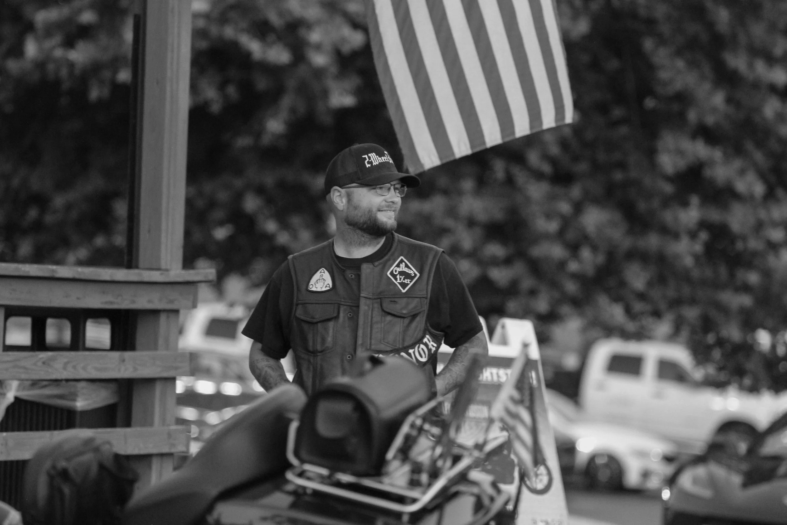 A smiling man with glasses, a beard, and tattoos on his arms, wearing a cap and a vest, standing outdoors near a motorcycle, with a striped flag hanging behind him and blurred vehicles in the background.