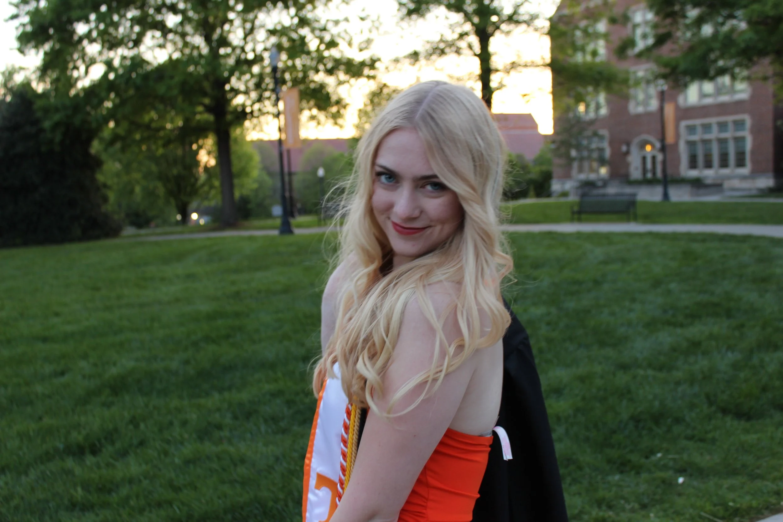 A young woman with long blonde hair and blue eyes, smiling, standing outdoors on a grassy area during sunset, with trees and brick buildings in the background.