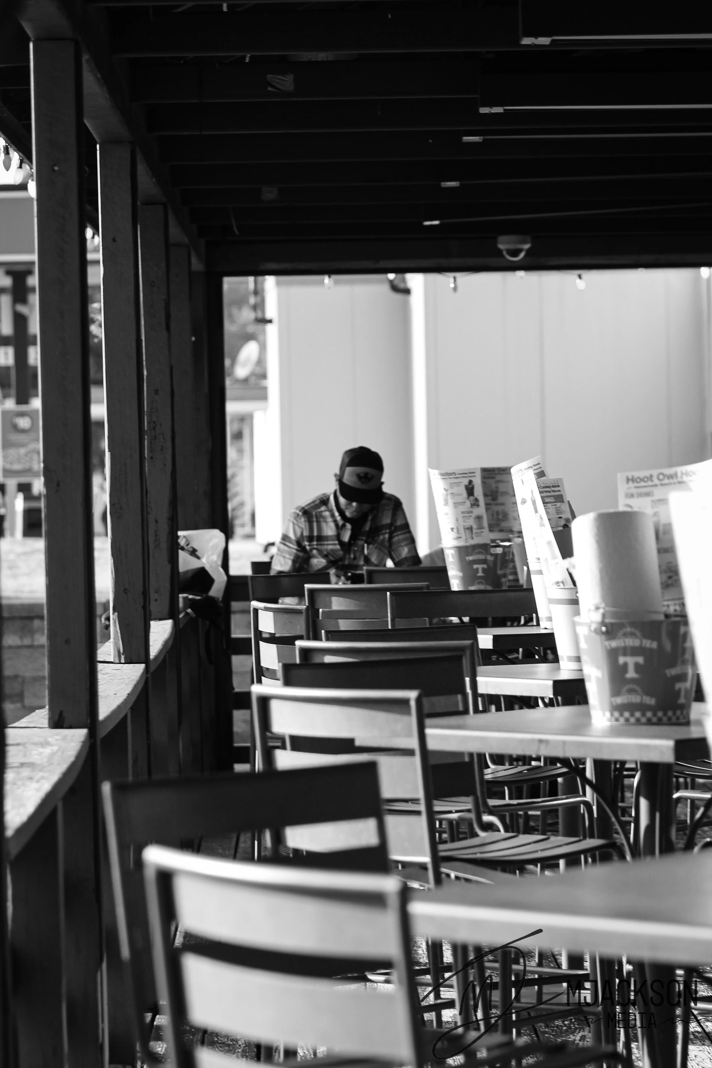 Empty outdoor seating area of a restaurant or cafe with a man sitting at a table, holding a phone, with drinks and menus on the tables, under a wooden pergola structure.