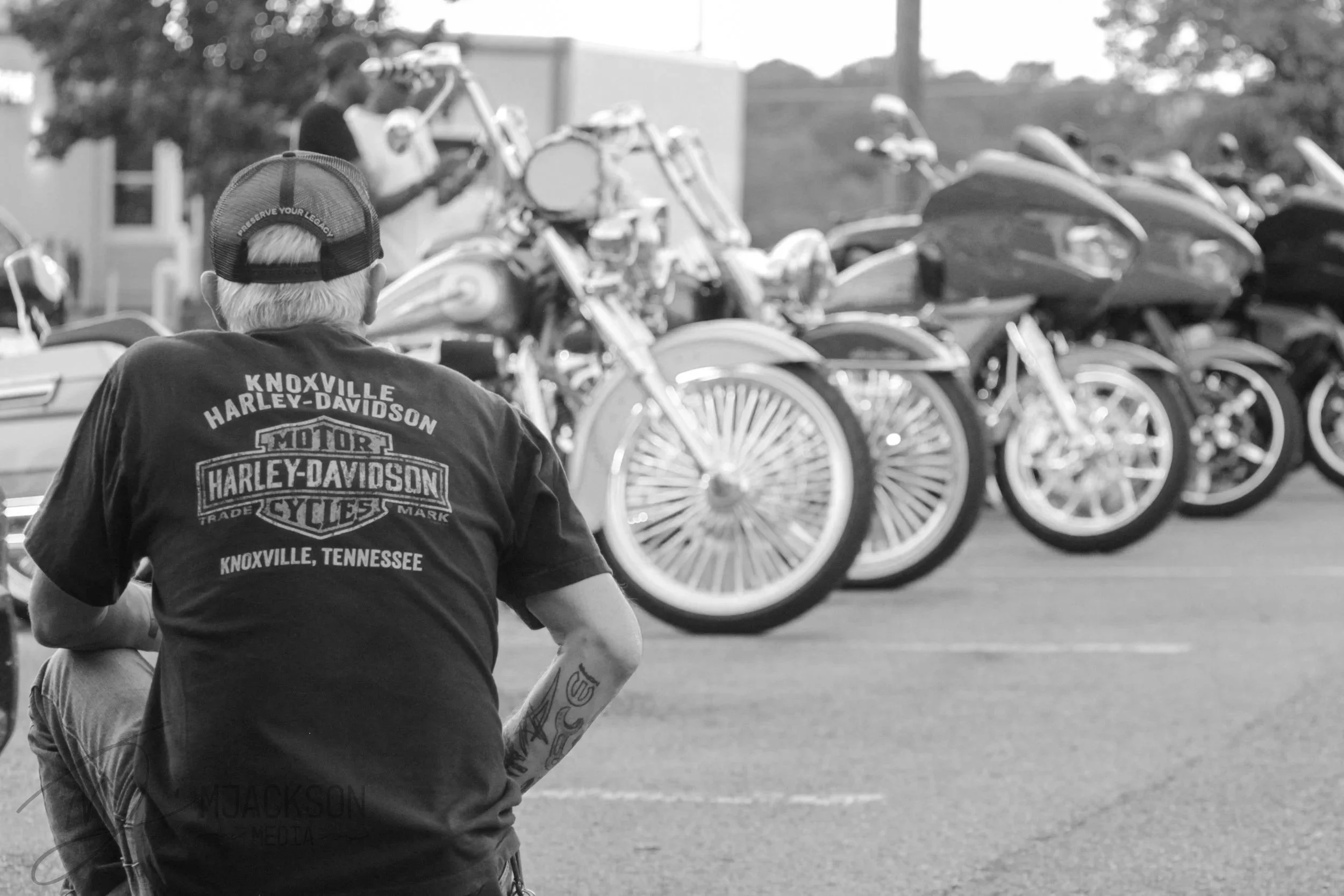 A person with gray hair wearing a black baseball cap with the words "Preserve Your Legacy" seated near a row of parked motorcycles, with the person's black t-shirt displaying "Knoxville Harley-Davidson" and a motorcycle logo.