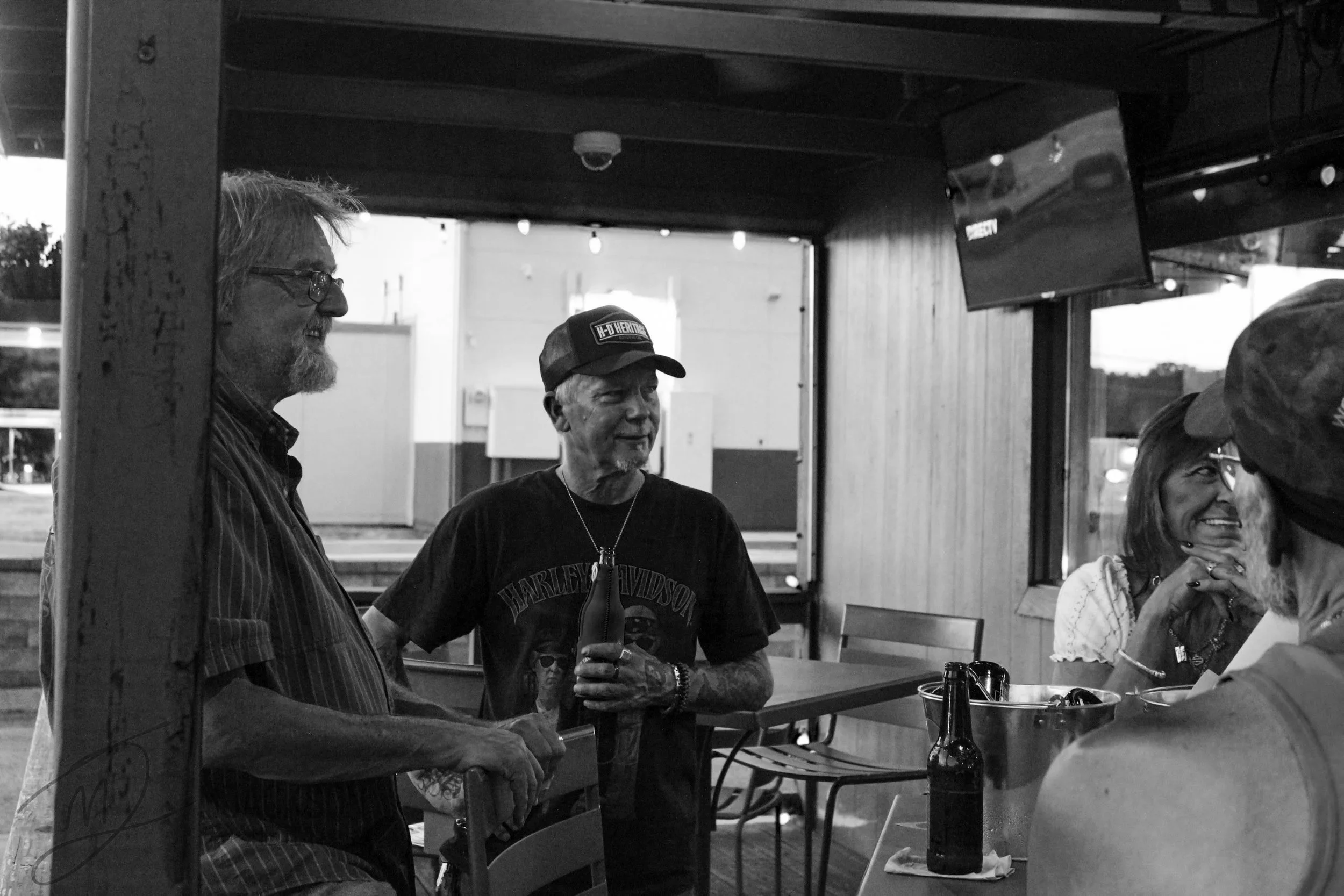 A group of five people gathered inside a casual venue, engaged in conversation and laughter. One man with glasses and a beard is smiling at another man wearing a Harley-Davidson cap, who is holding a drink. A woman with long hair is smiling and holdi