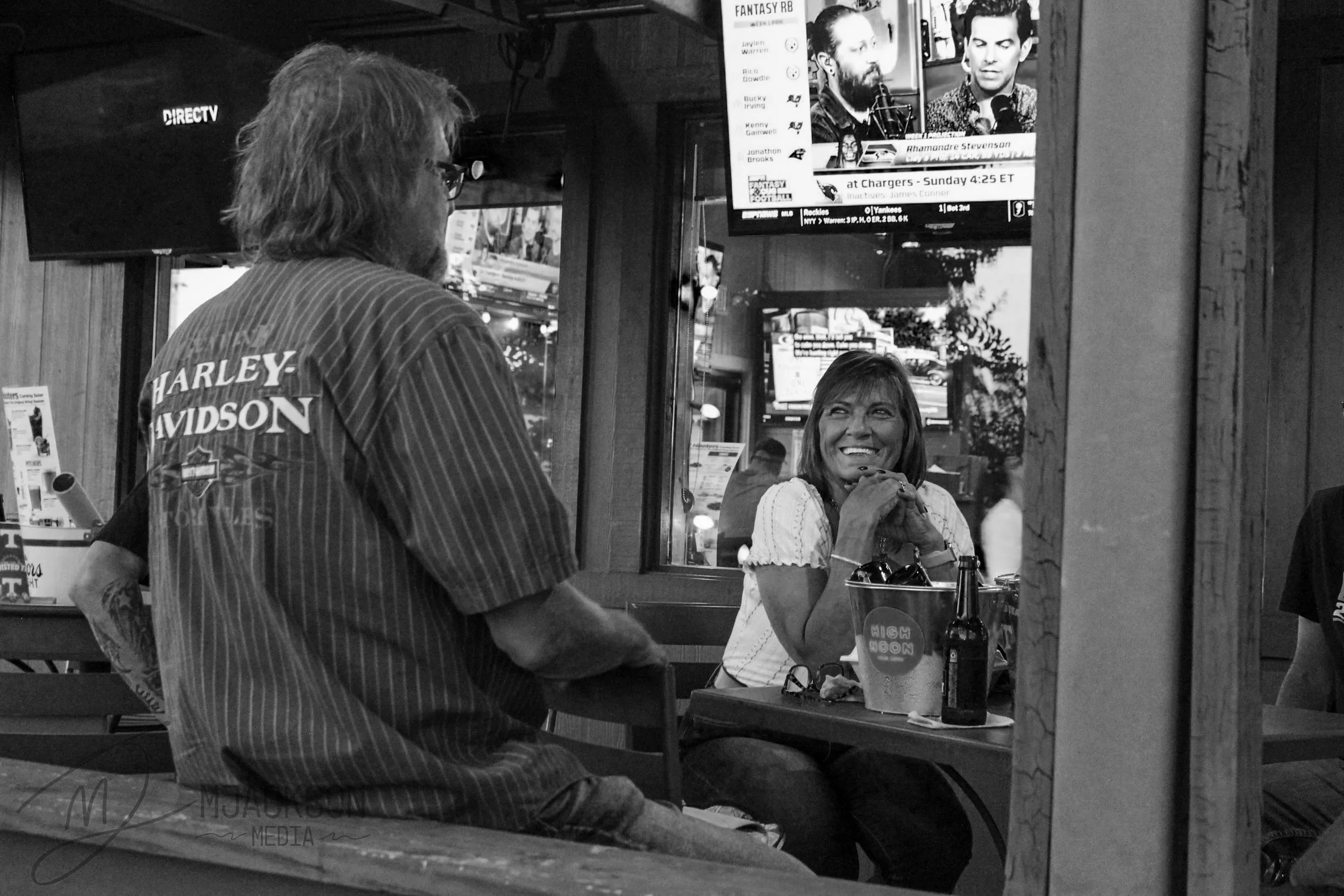 A man with long hair and glasses wearing a shirt with 'Harley Davidson' on the back is standing in front of a woman seated at a bar or counter. The woman is smiling, resting her chin on her hands, and looking at the man. There are multiple TV screens