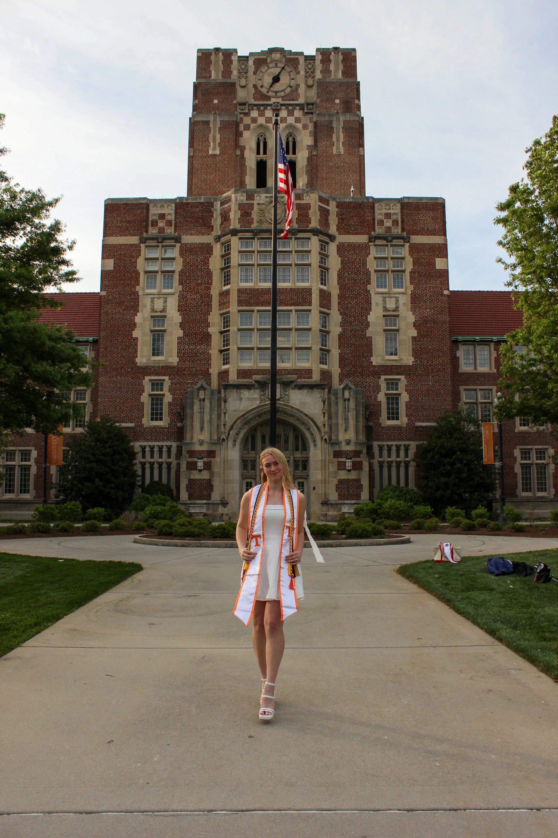 A young woman in a white dress and high heels standing on a sidewalk in front of a historic brick building with a clock tower, surrounded by trees and greenery, during daytime.