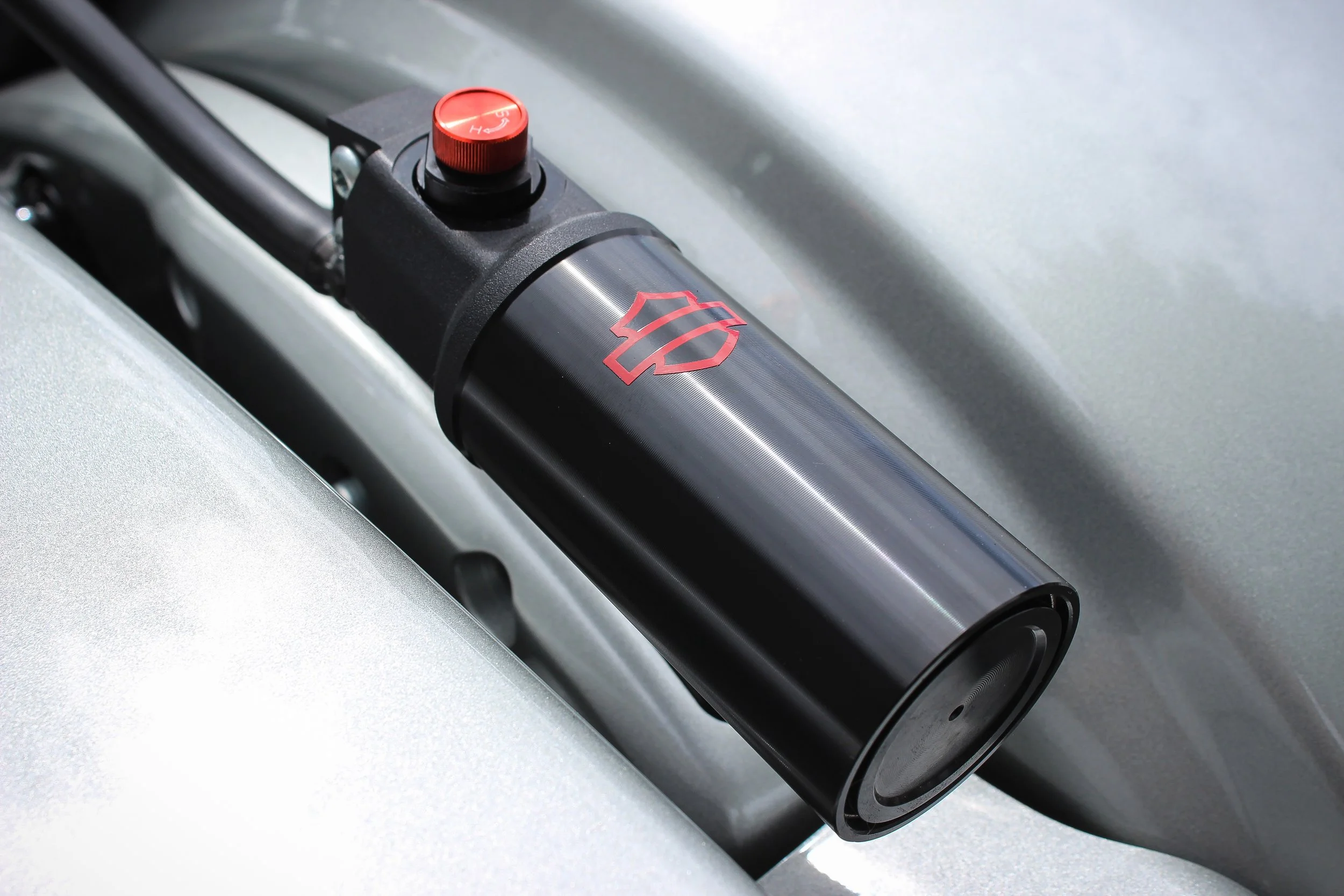 Close-up of a hydraulic damper with a red adjustment knob and a red logo on a silver surface.