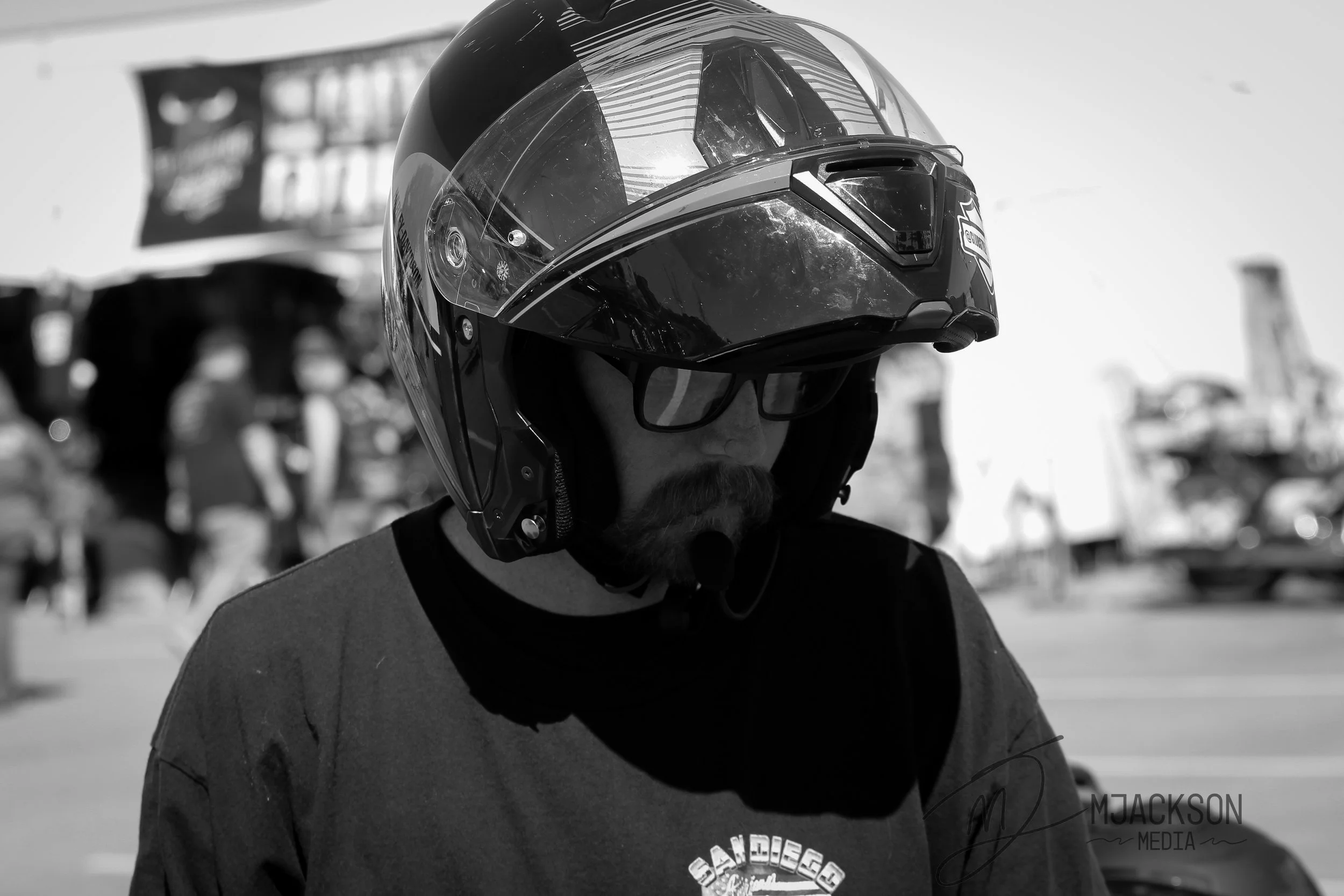 A person wearing a motorcycle helmet and sunglasses, standing outdoors in a sunny environment with rides and structures in the background.