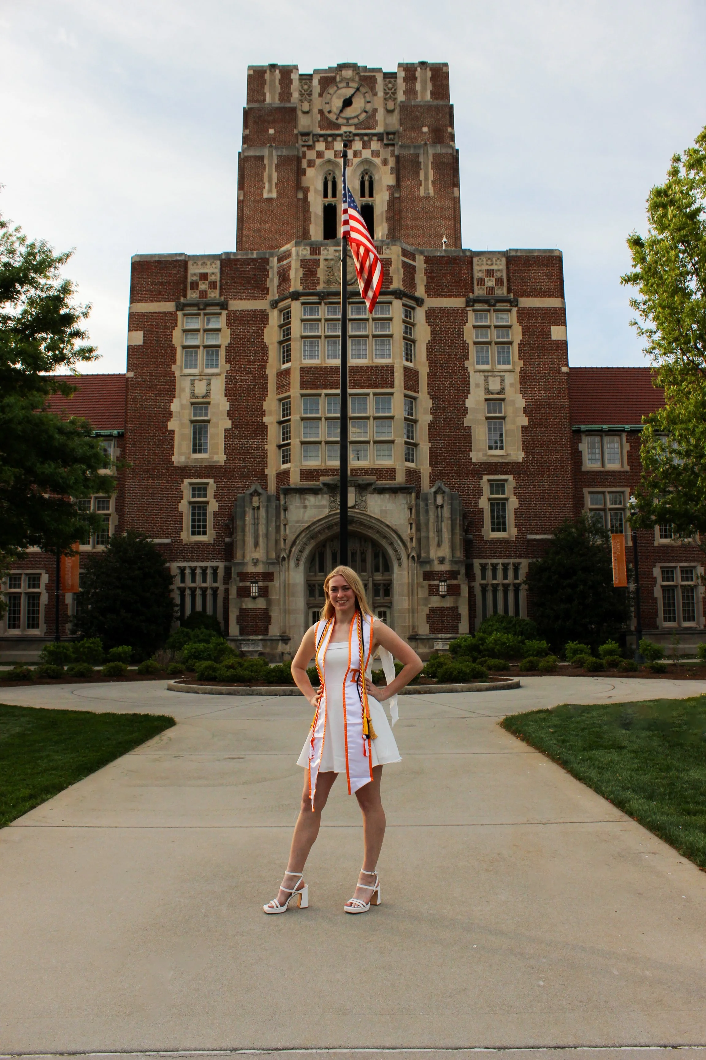 A woman in a white dress and high heels standing confidently on a university campus with a large brick clock tower in the background and an American flag in front of the tower.