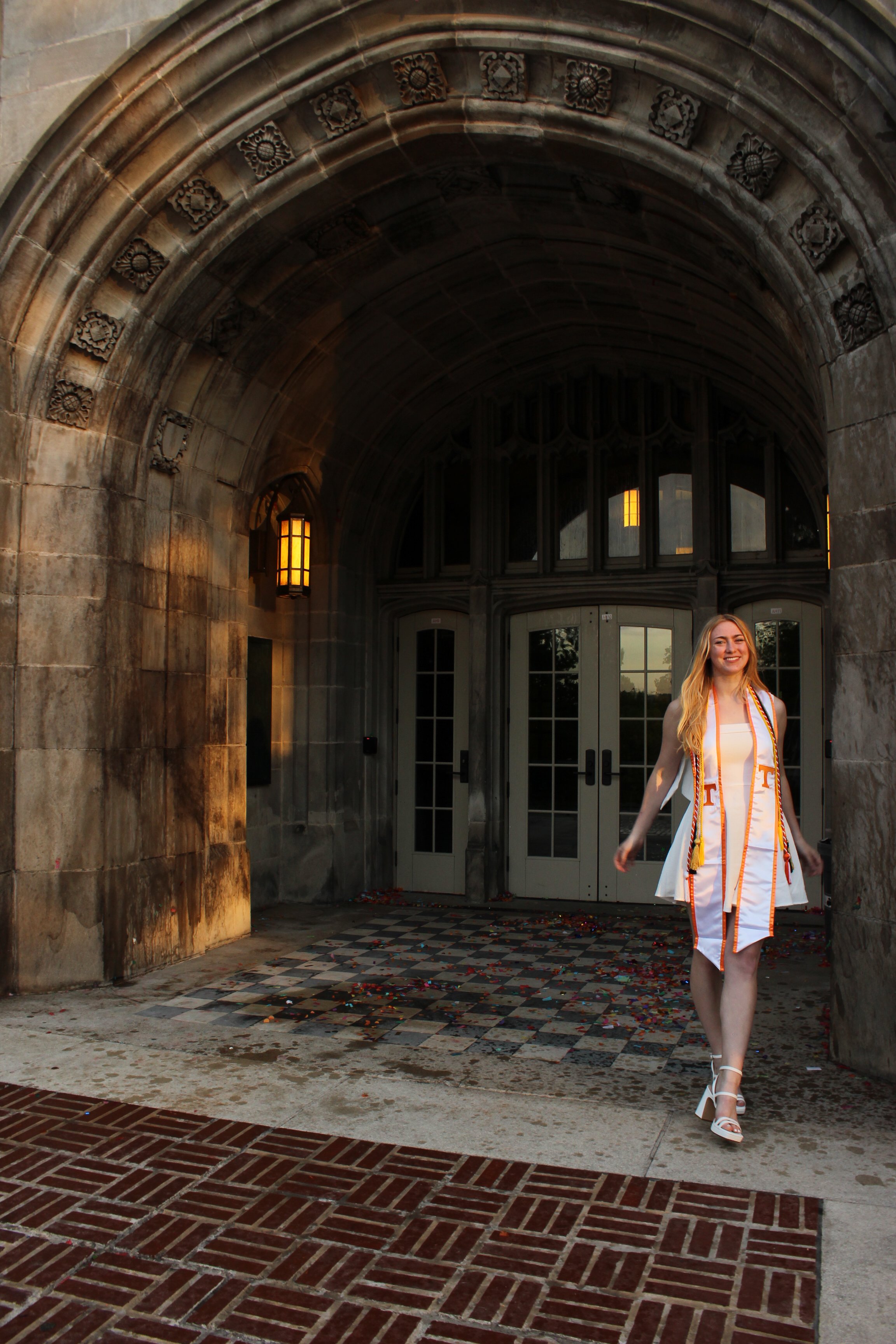 A young woman in a white gown with orange accents and white high heels walking under a stone archway on a checkered tile floor, celebrating graduation, with confetti on the ground and a building with large windows in the background.