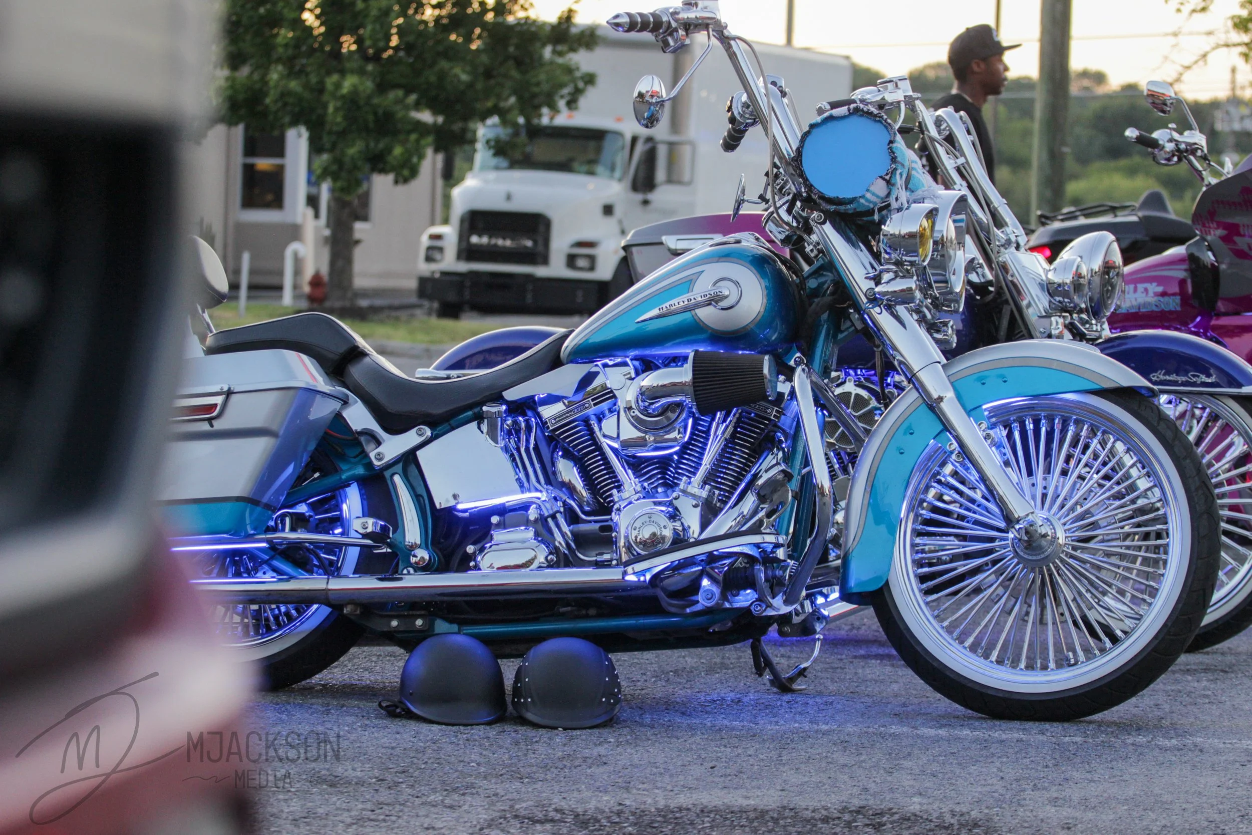 A row of parked Harley Davidson motorcycles, including a prominent blue and chrome bike in the foreground with black helmets placed on the ground nearby, set outdoors during sunset with a man standing in the background.
