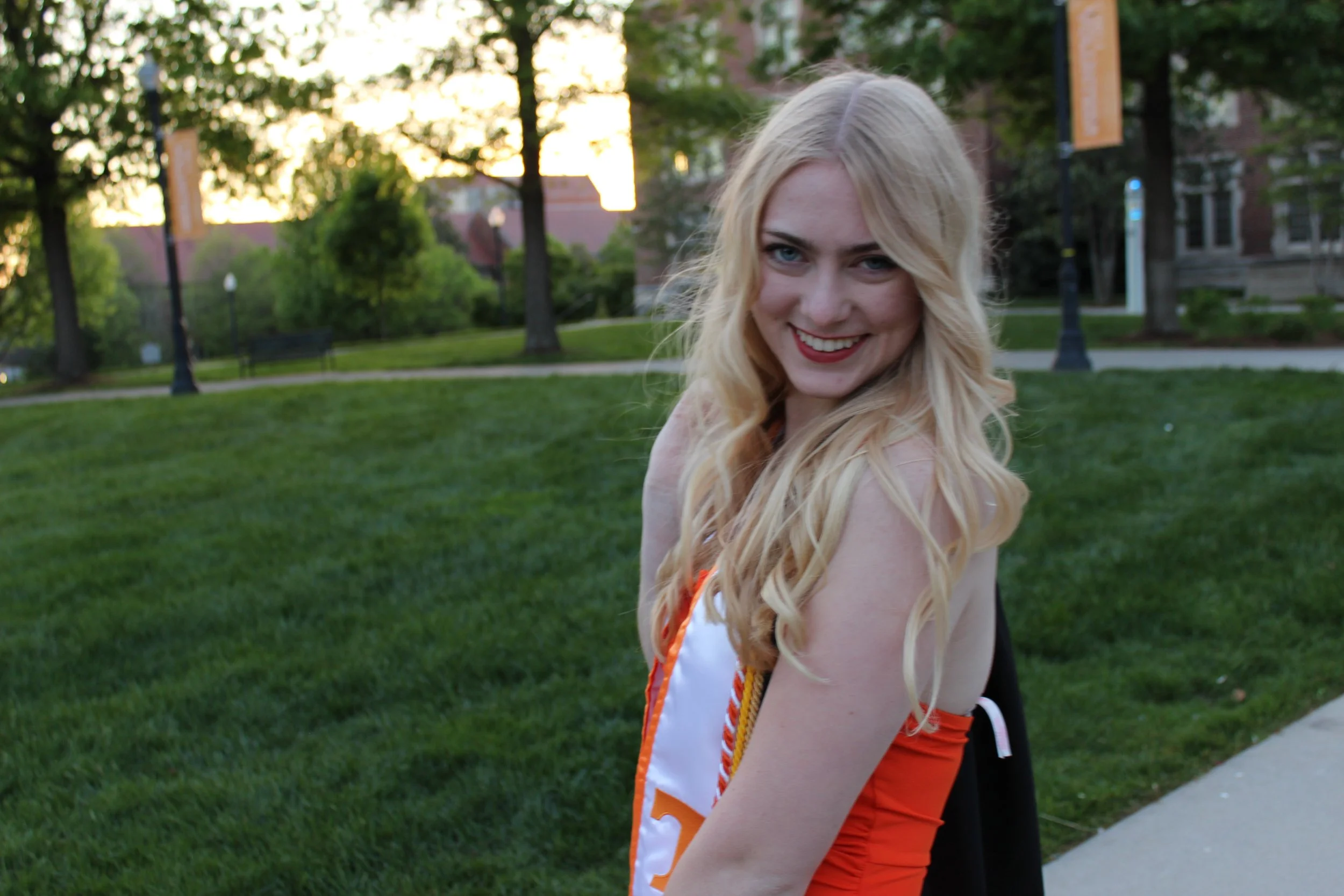 A young woman with long blonde hair smiling outdoors in a park during sunset, wearing a cheerleading uniform with orange, white, and black colors.
