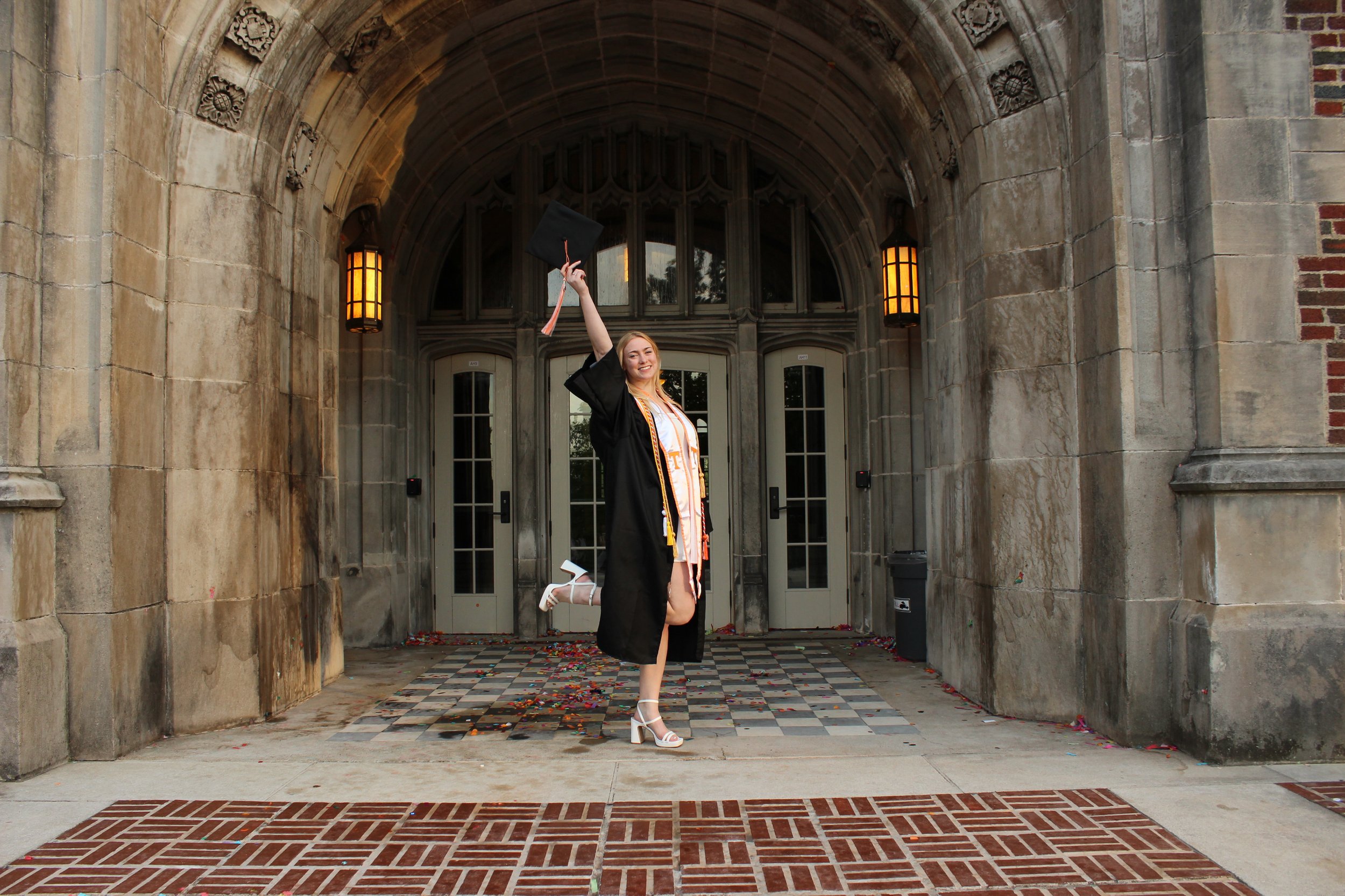 A woman in a graduation gown and cap stands in an arched stone entrance holding her cap in the air and smiling, with confetti on the ground.