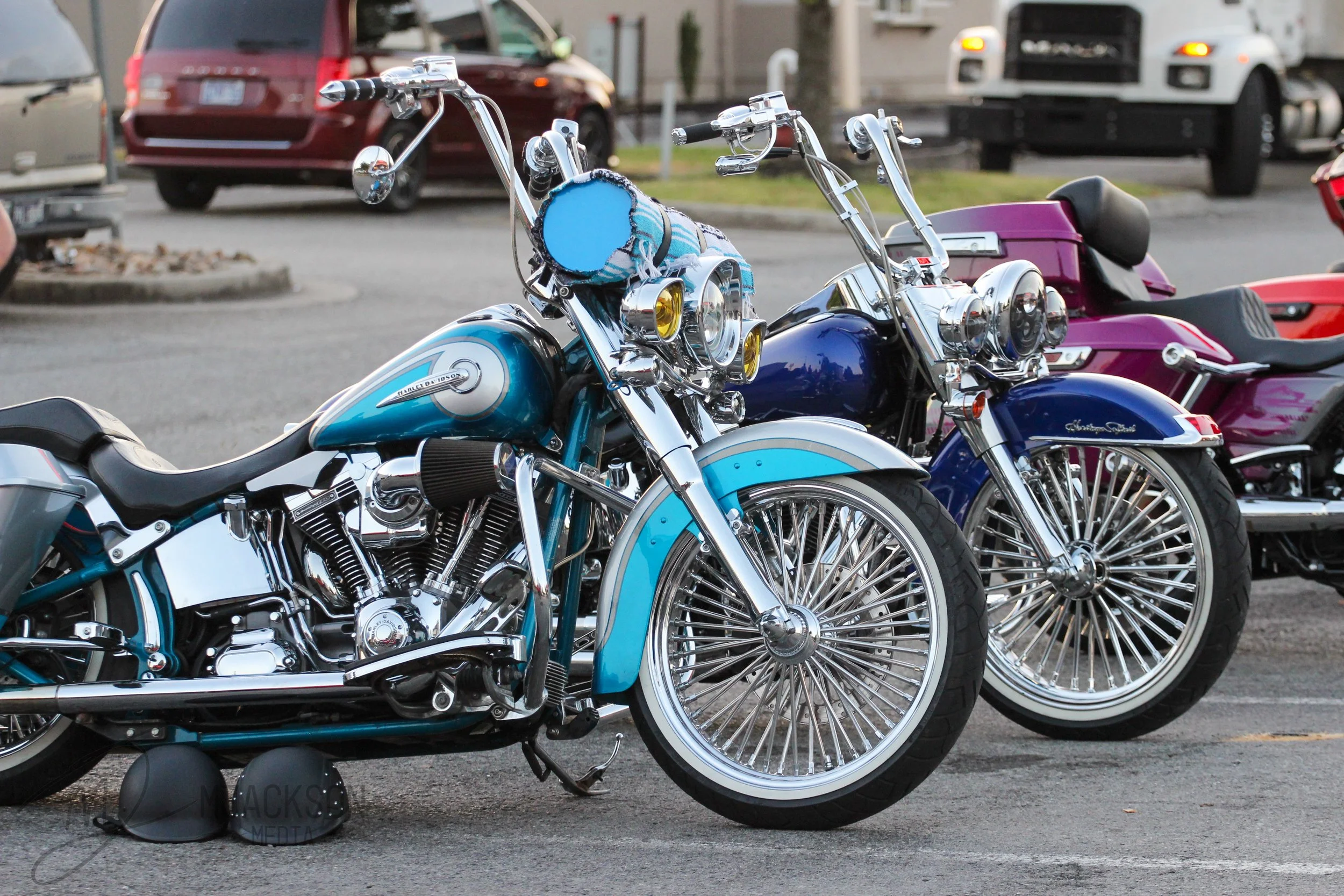 A row of parked motorcycles, including a blue Harley Davidson with a fringed saddlebag, in a parking lot with cars and trucks in the background.