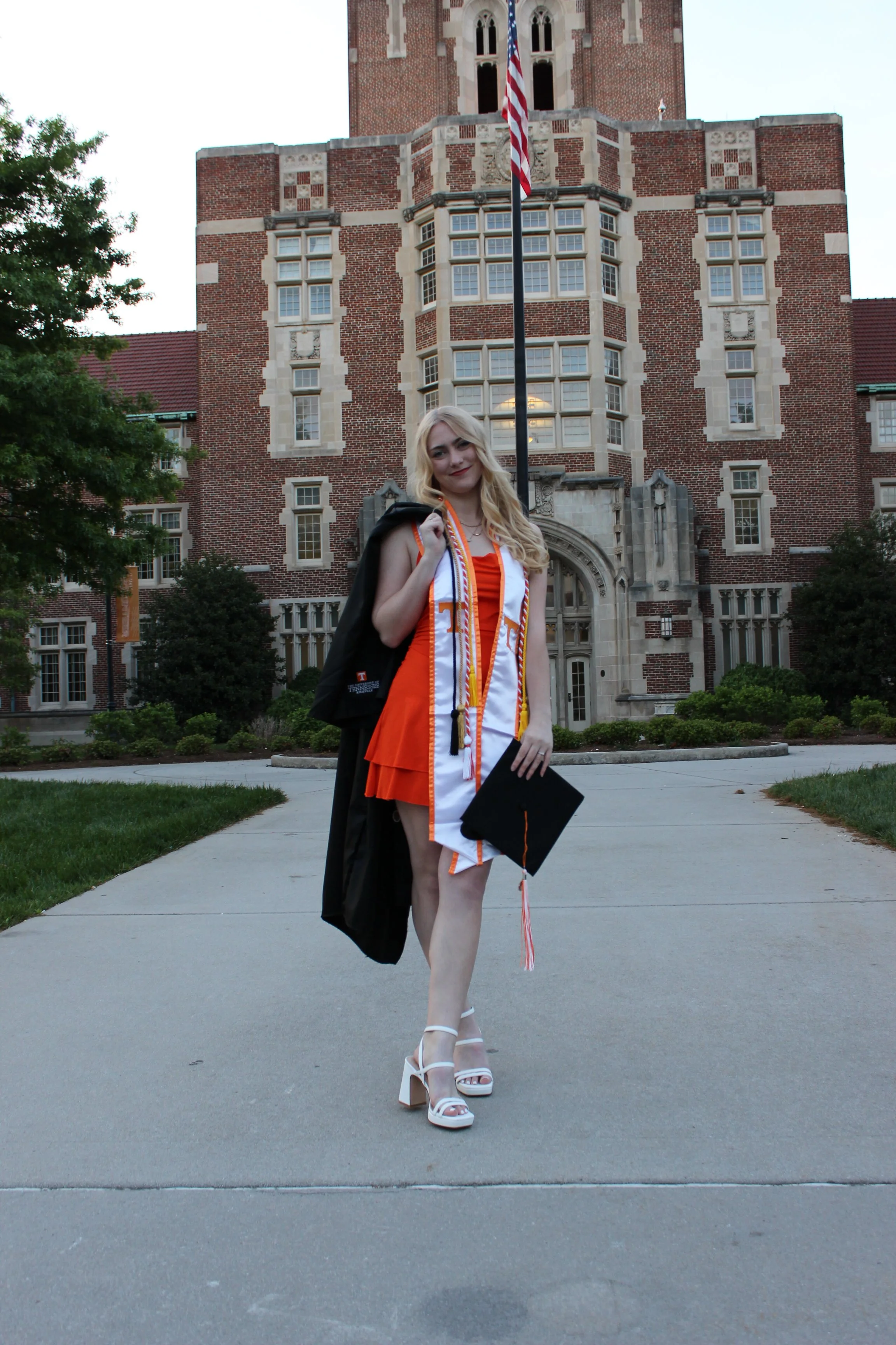 A young woman in an orange dress with white high heels, doctor’s garb around waist, and honor cords with tassels, standing in front of a brick building with gothic architecture, holding a black cap, possibly at a graduation ceremony.