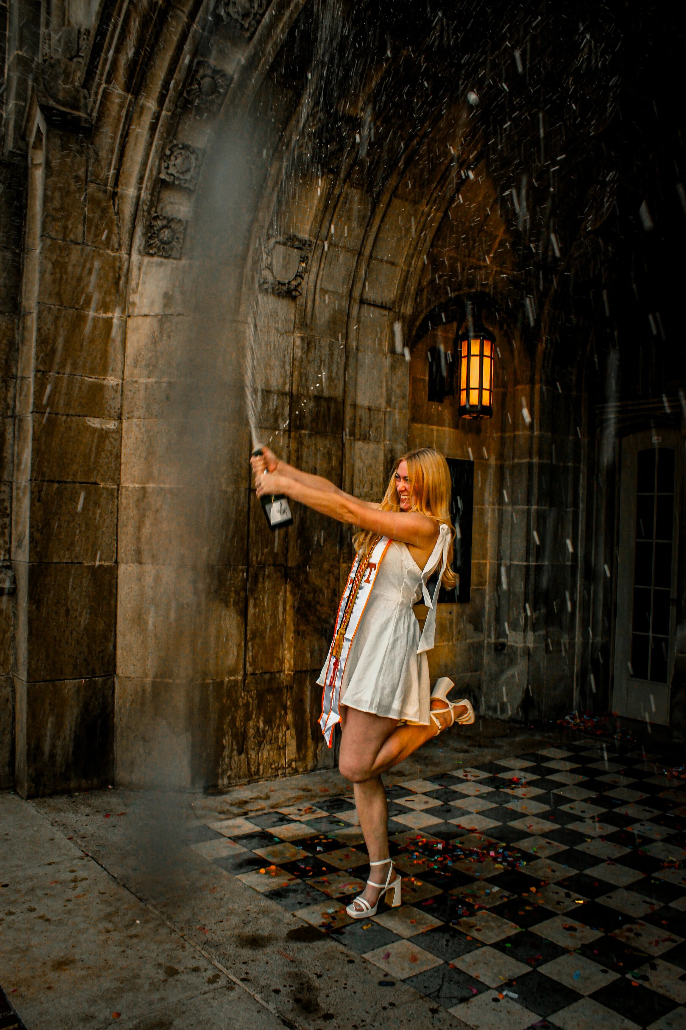 A woman in a white dress and high heels is celebrating by popping a champagne bottle in a doorway with stone walls and a lantern. Confetti is scattered on the ground.