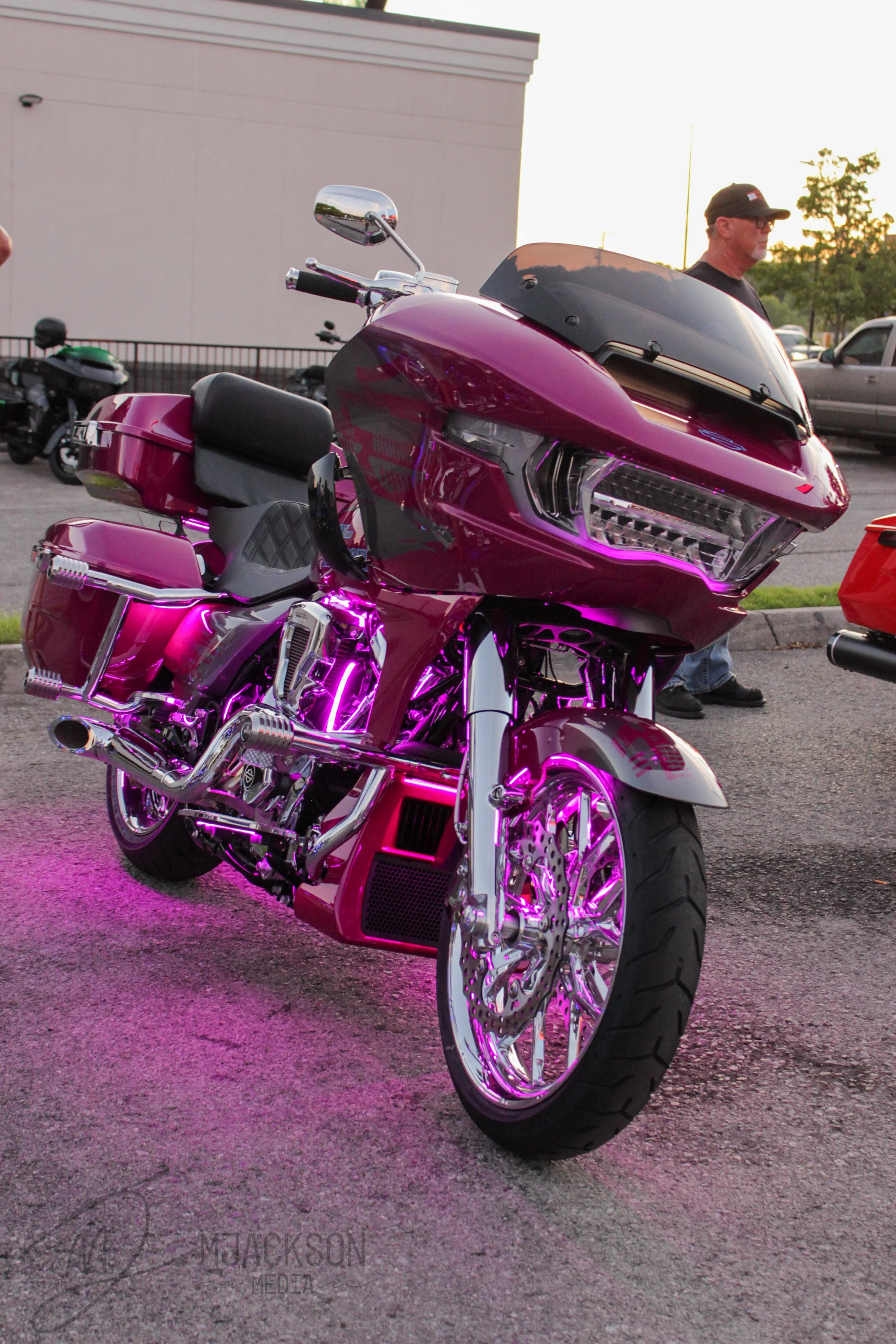 A shiny purple motorcycle with neon purple lighting accents parked outdoors, with a man wearing glasses and a cap standing nearby during sunset.