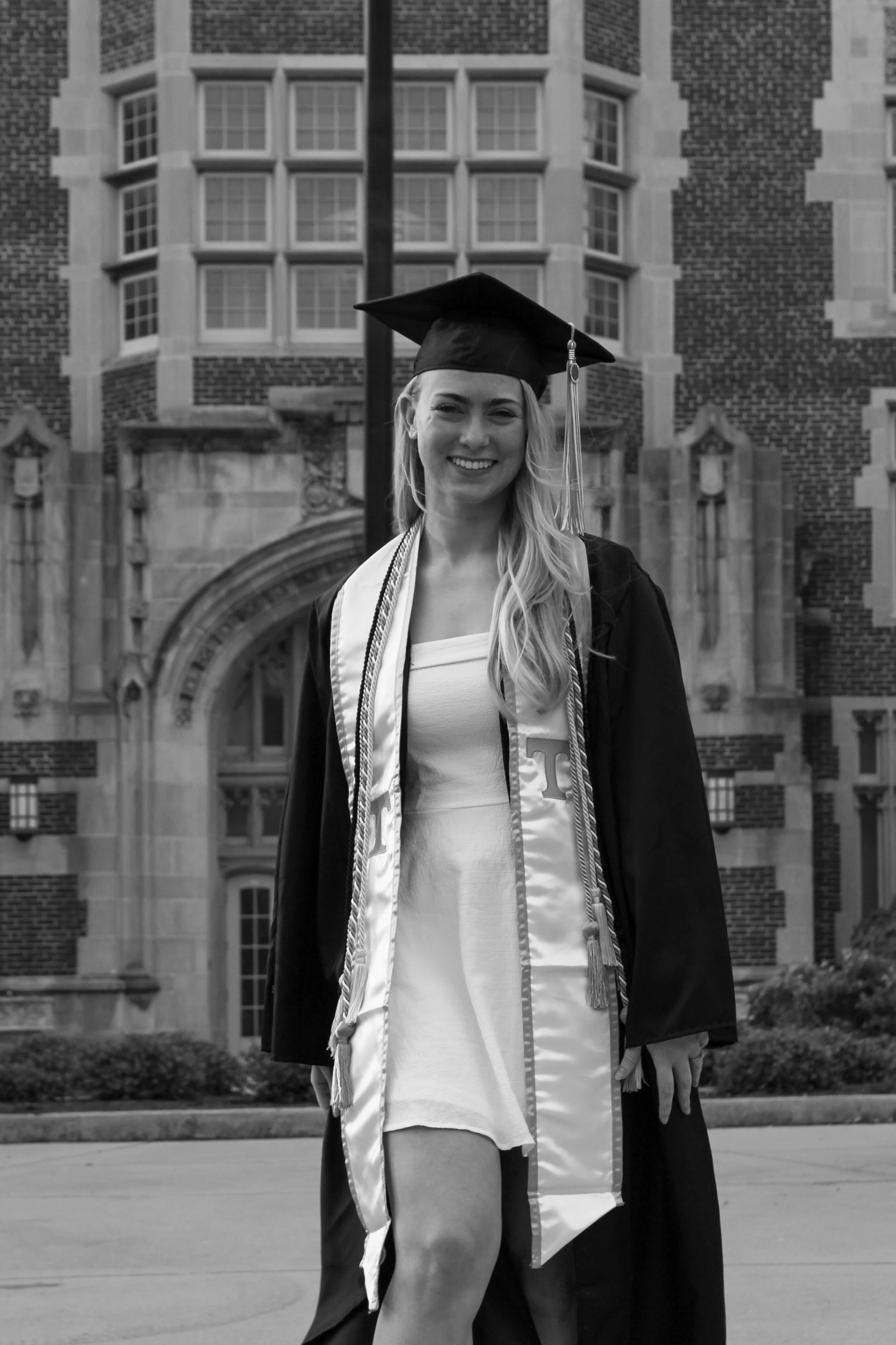 Smiling young woman in graduation cap and gown standing in front of university building.