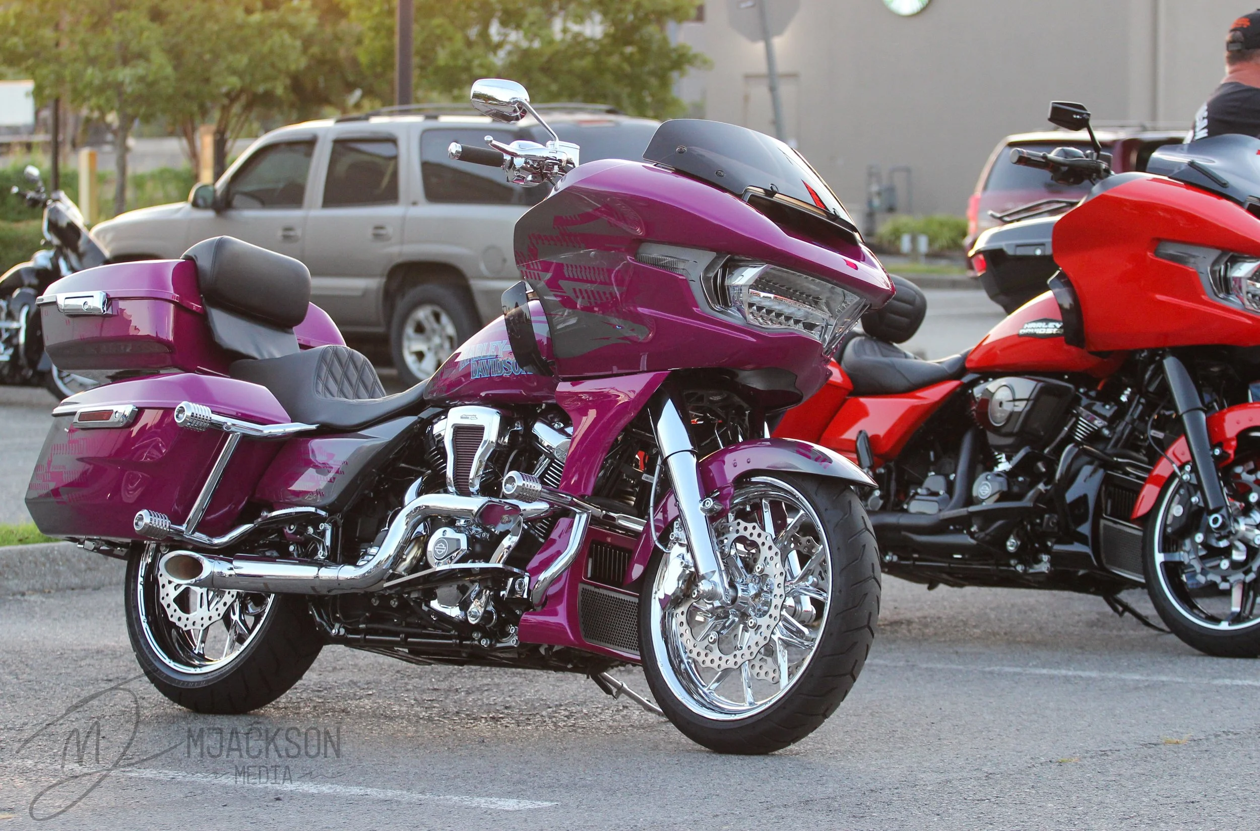 Multiple motorcycles, including a purple Harley-Davidson touring bike and a red motorcycle, parked outdoors with cars and trees in the background.