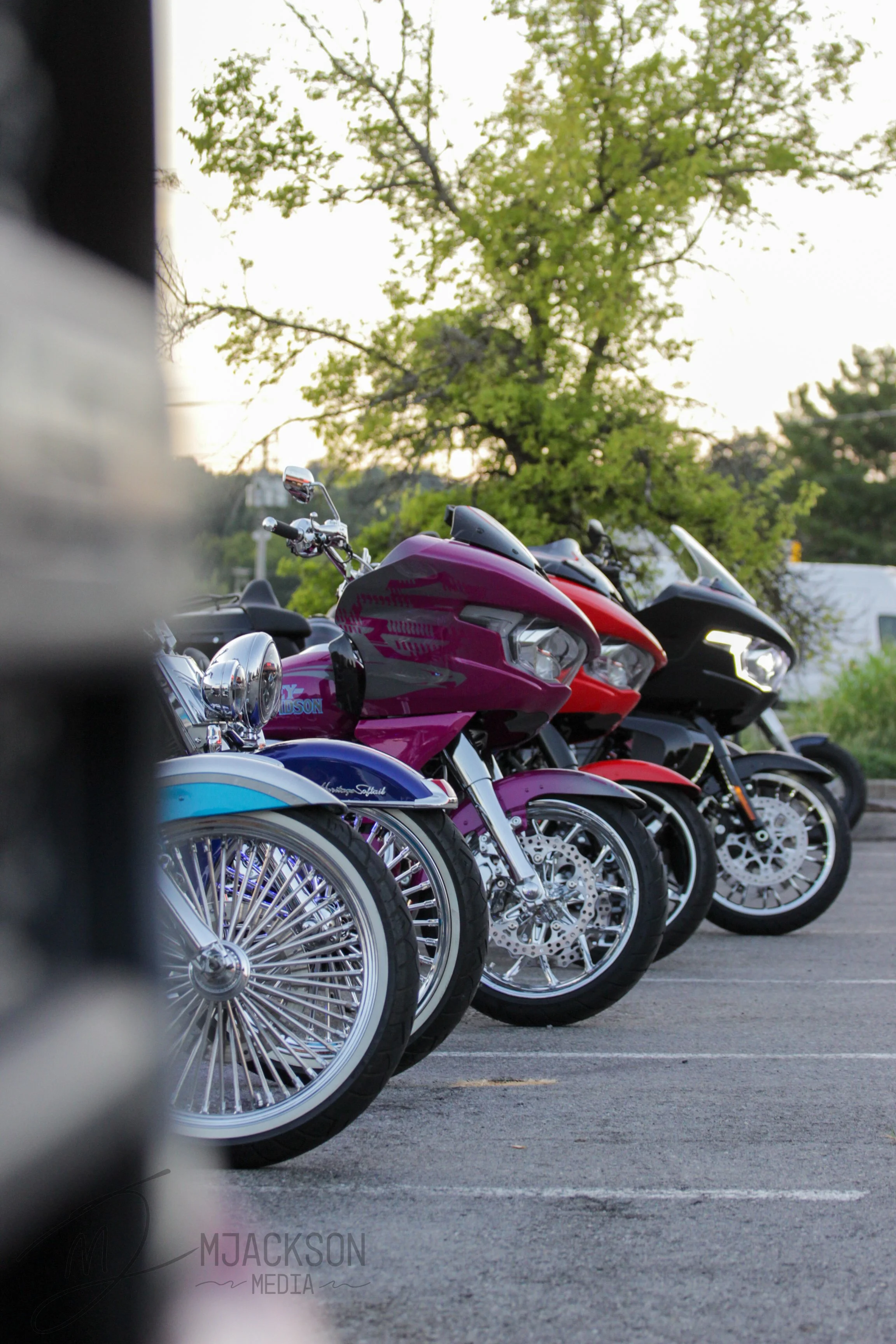 Multiple motorcycles parked in a row in an outdoor parking lot, with green trees and a clear sky in the background.