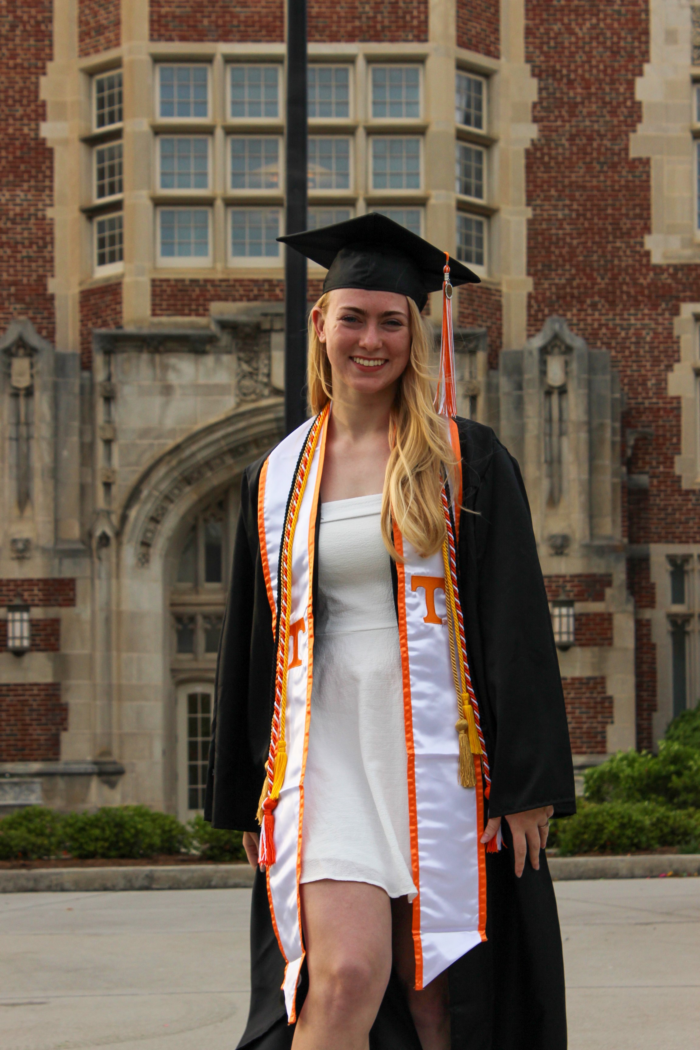 A smiling young woman in a black graduation cap and gown with orange and white honors cords, standing in front of a brick university building.