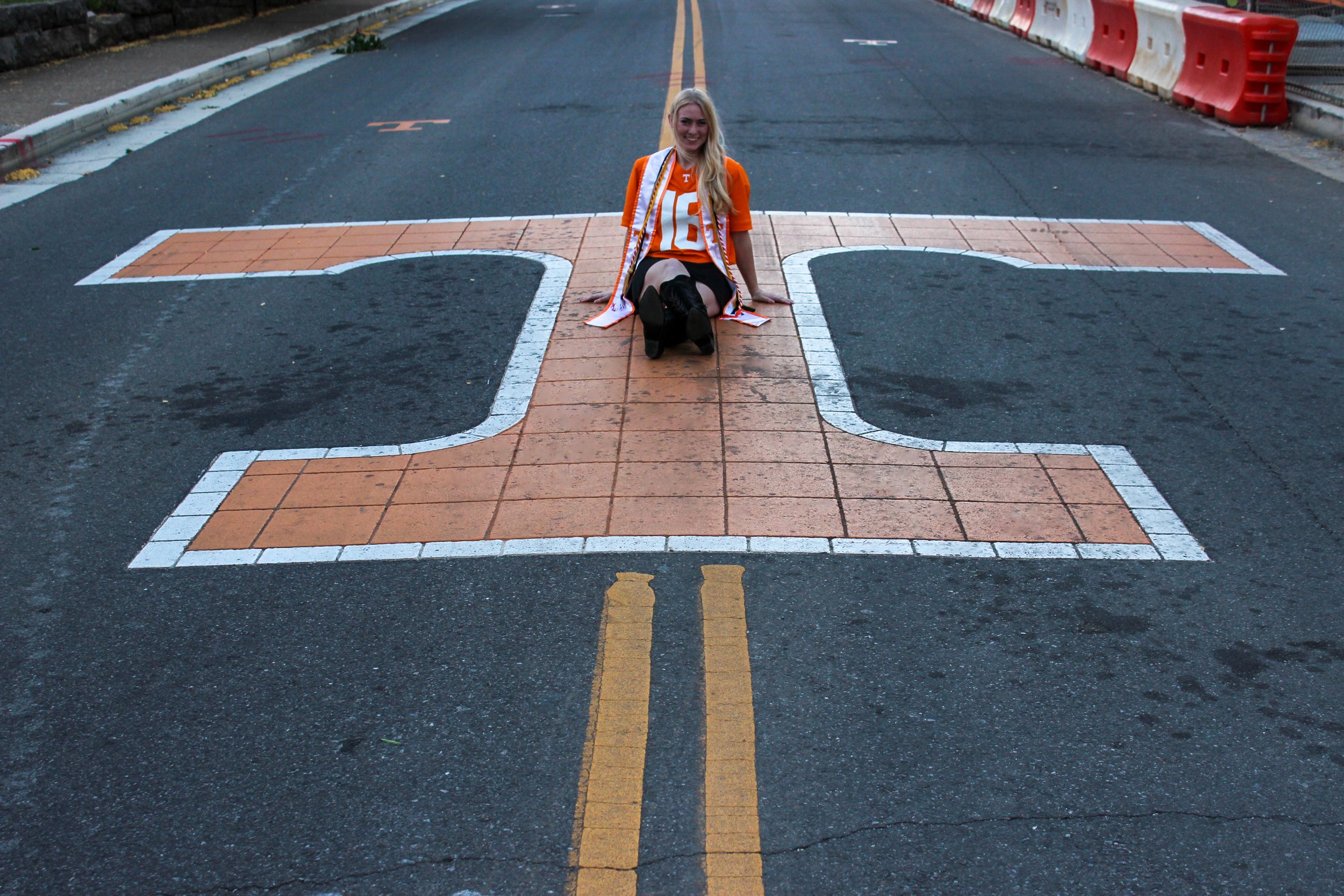 A young woman with long blonde hair, wearing an orange University of Tennessee jersey, sits on a crosswalk painted on an asphalt street. She is smiling and facing the camera, with construction barriers visible along the side of the road.