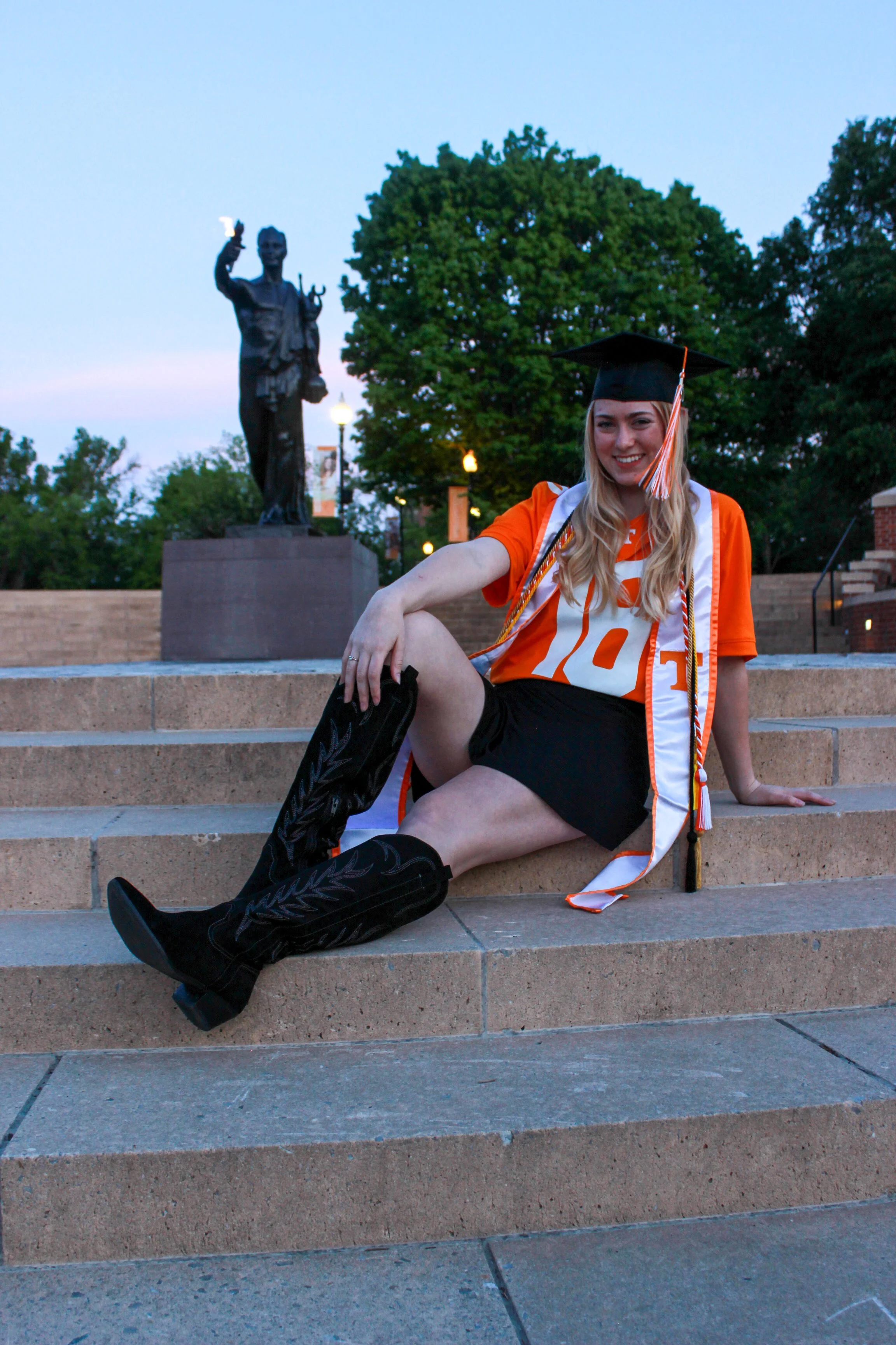 A young woman in a graduation cap and gown sitting on stairs outdoors, smiling, with a statue in the background and trees around during dusk.