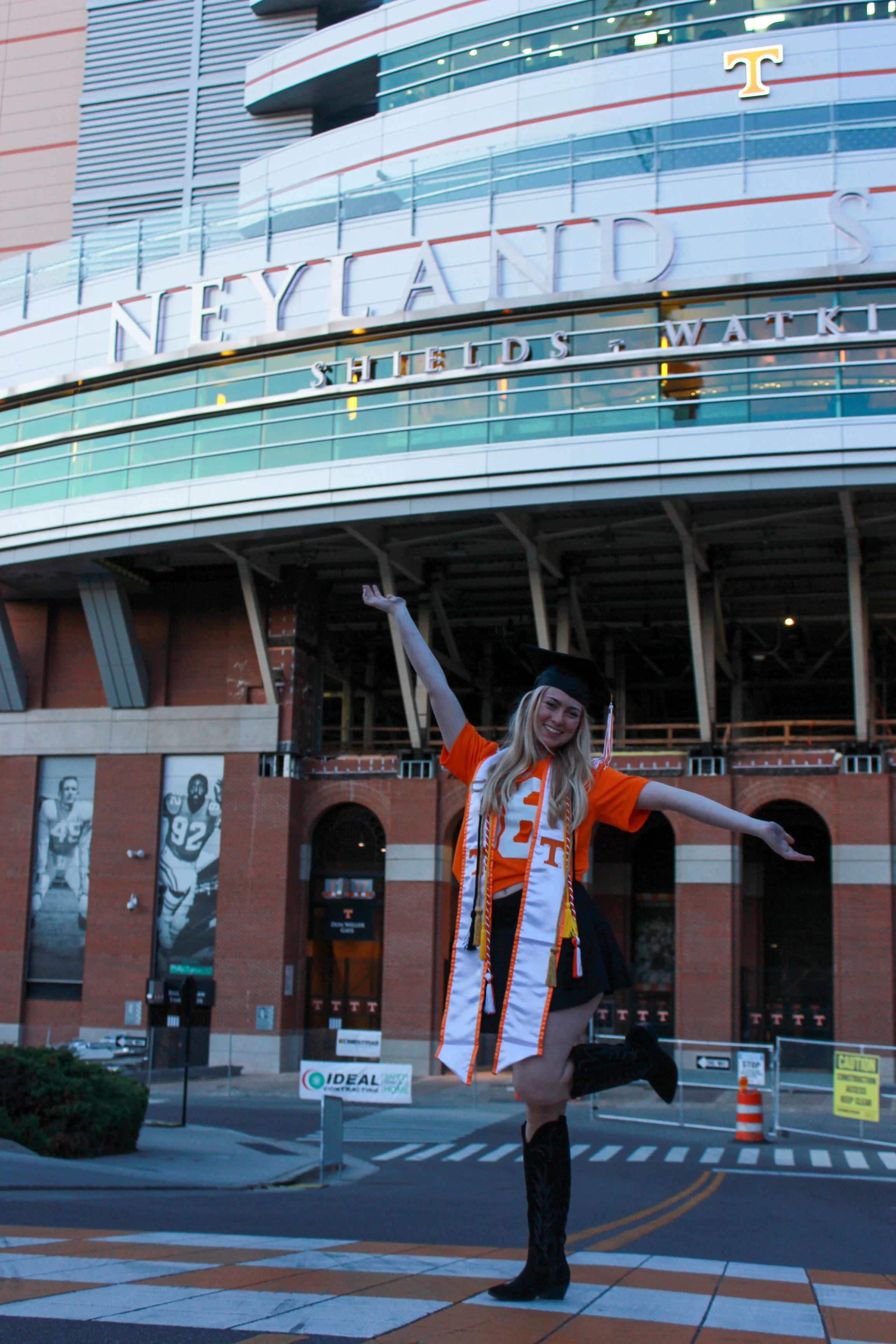 A young woman dressed in an orange and white dress with the Tennessee Volunteers logo, black thigh-high boots, and a graduation cap, celebrating in front of the Neyland Stadium in Knoxville, Tennessee, with her arm raised and smiling.