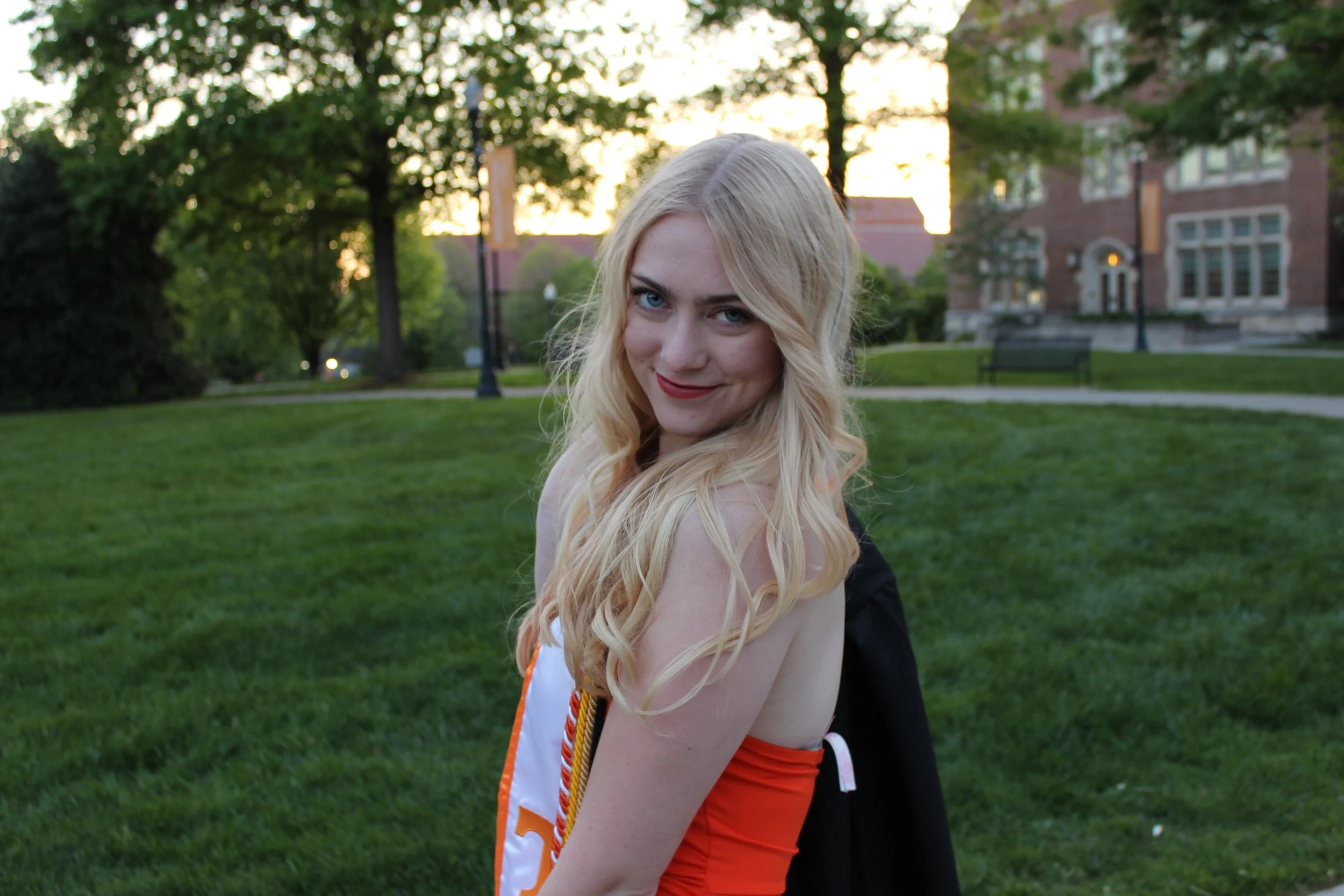 A young woman with long blonde hair and blue eyes smiling in a park with green grass, trees, and a brick building in the background during sunset.